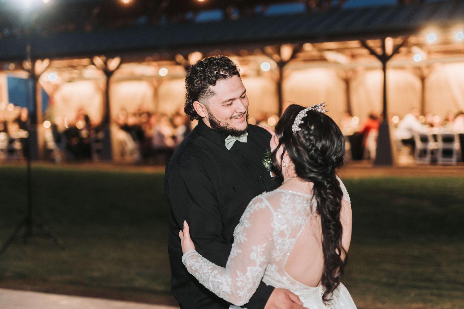 A couple shares a tender moment in formal wedding attire against a warmly lit outdoor pavilion at dusk.