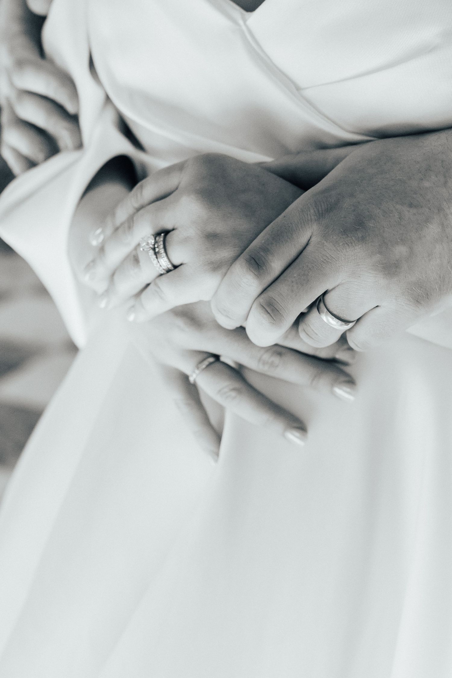 Close-up black and white photo of two hands with wedding rings resting on white fabric.