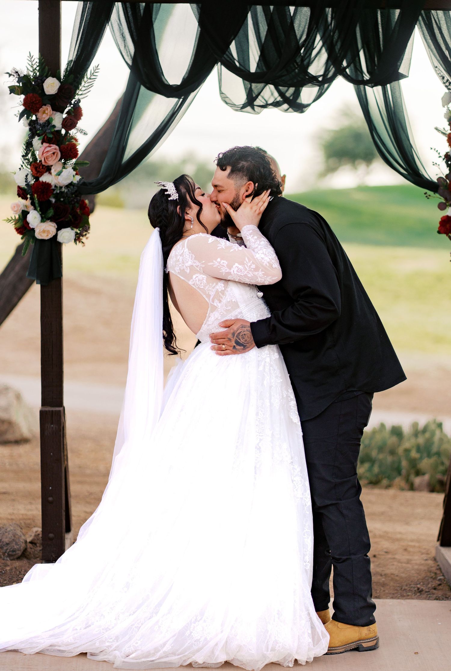 A couple shares their first kiss at a wedding ceremony under black fabric draping and floral arch decorations outdoors.