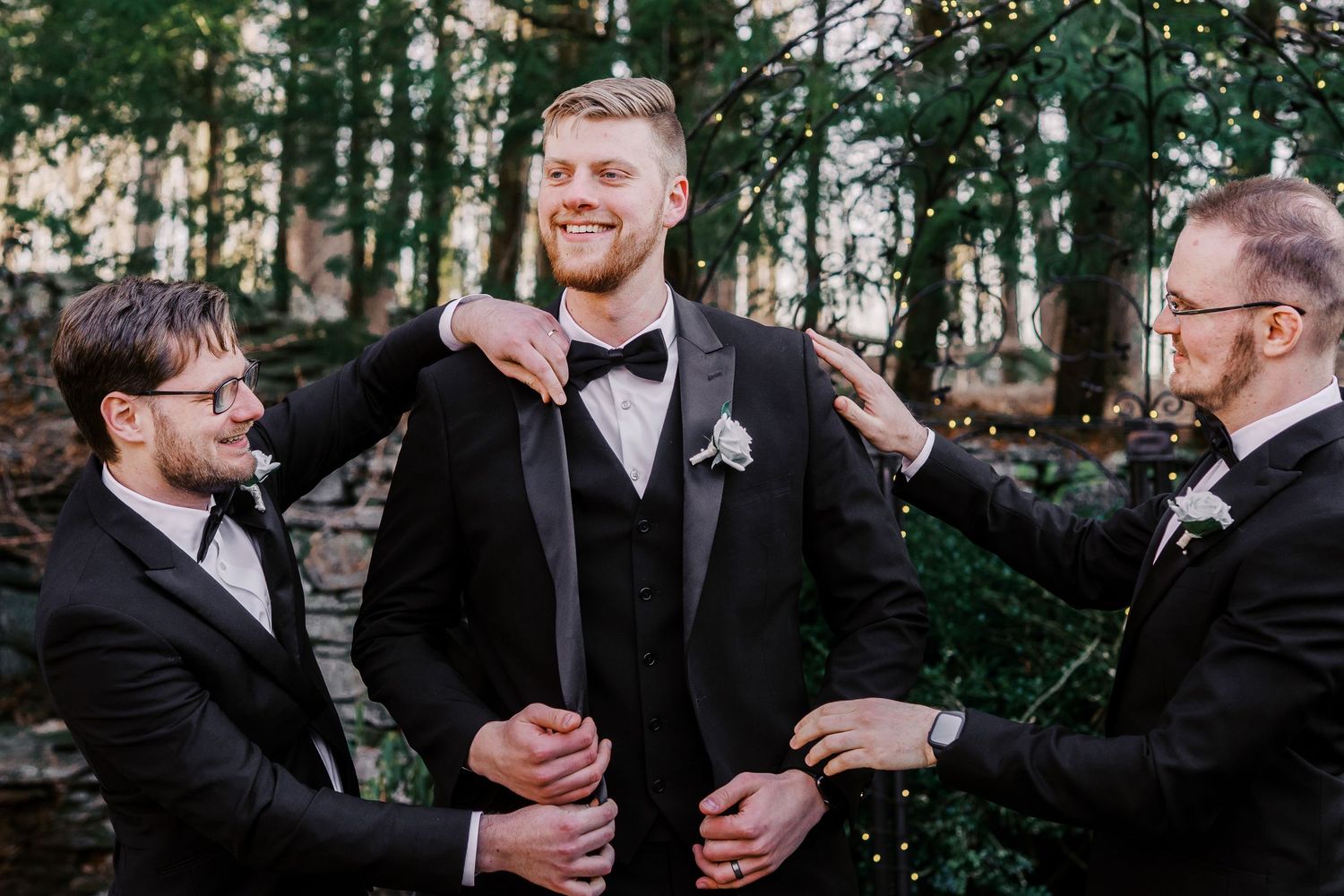 Groomsmen in black tuxedos and bow ties sharing a lighthearted moment in a forested outdoor setting.