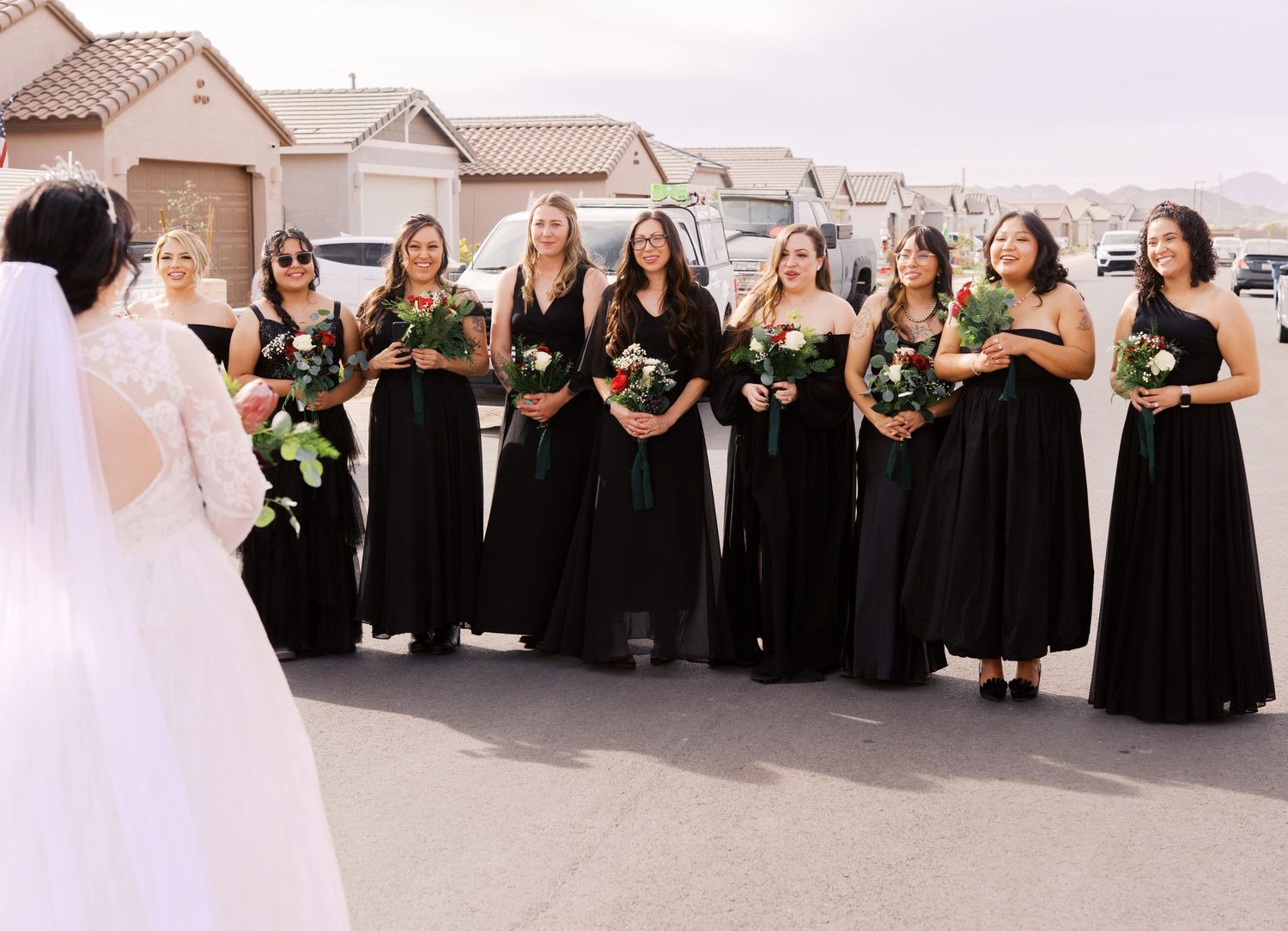 A wedding party in long black bridesmaid dresses holding floral bouquets stands together outdoors near residential homes.