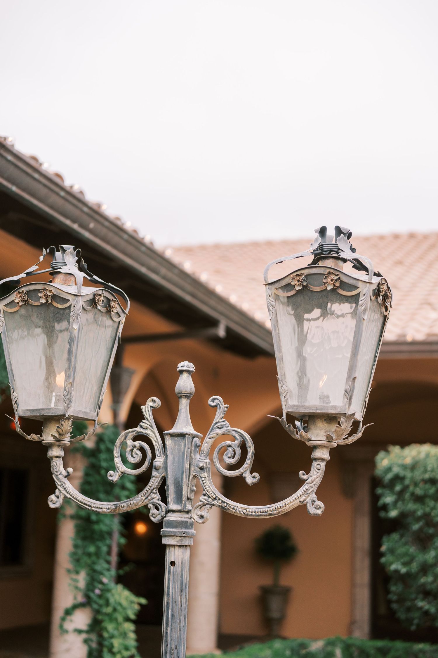 Ornate vintage-style double lamp post with decorative scrollwork and glass lanterns mounted against a rustic building exterior.