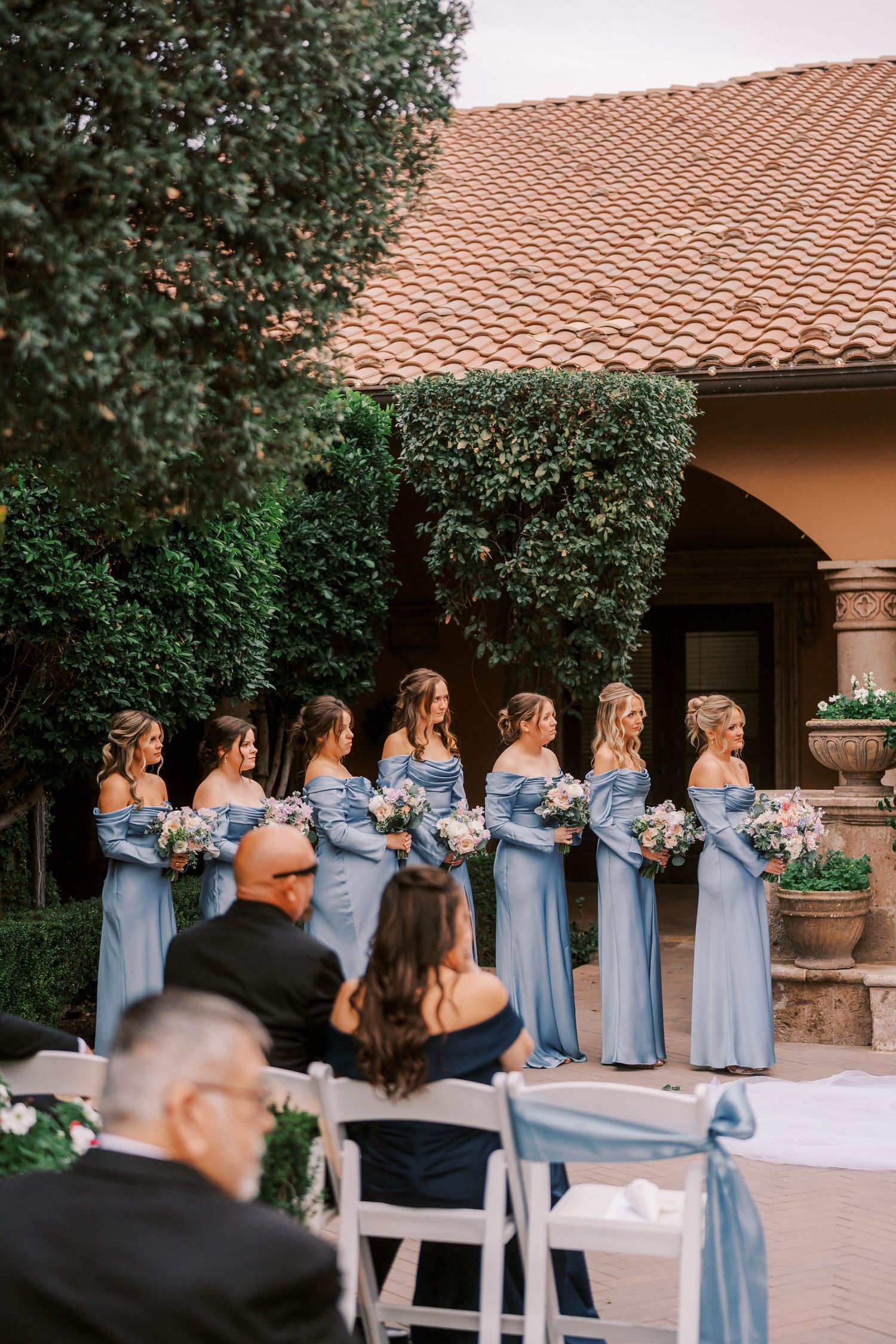Bridesmaids in light blue dresses stand in a line at an outdoor wedding venue with terracotta roof tiles and greenery.