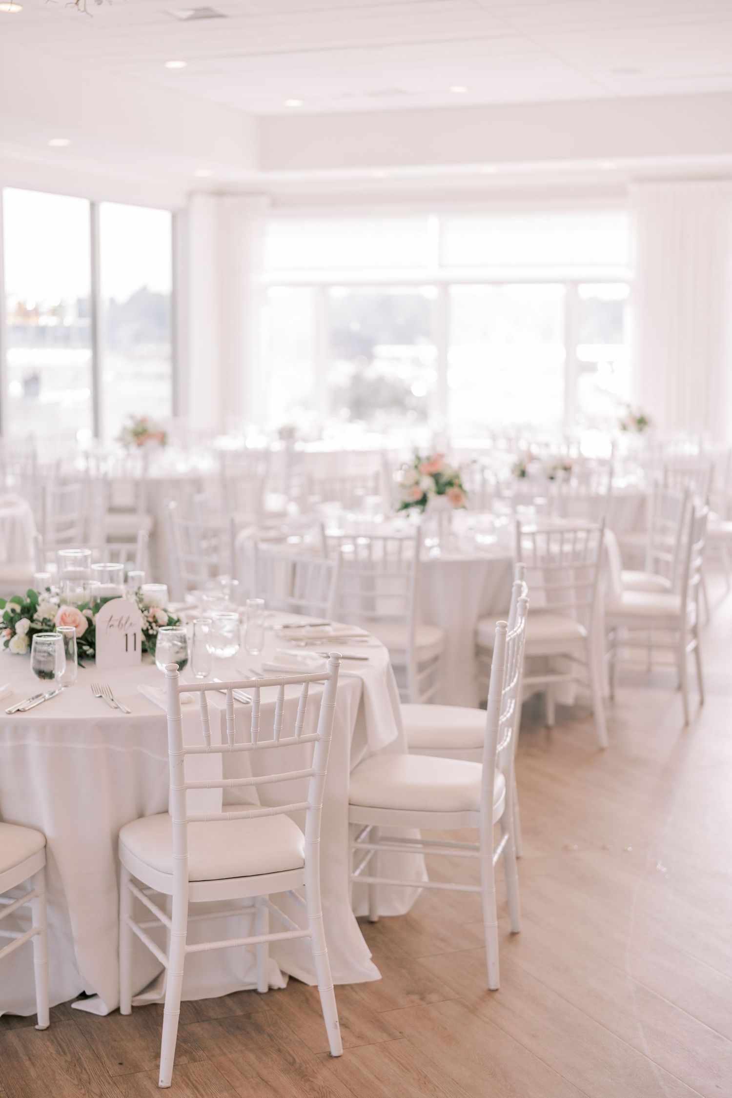 Elegant white wedding reception venue with Chiavari chairs, round tables, and soft floral centerpieces in natural light.
