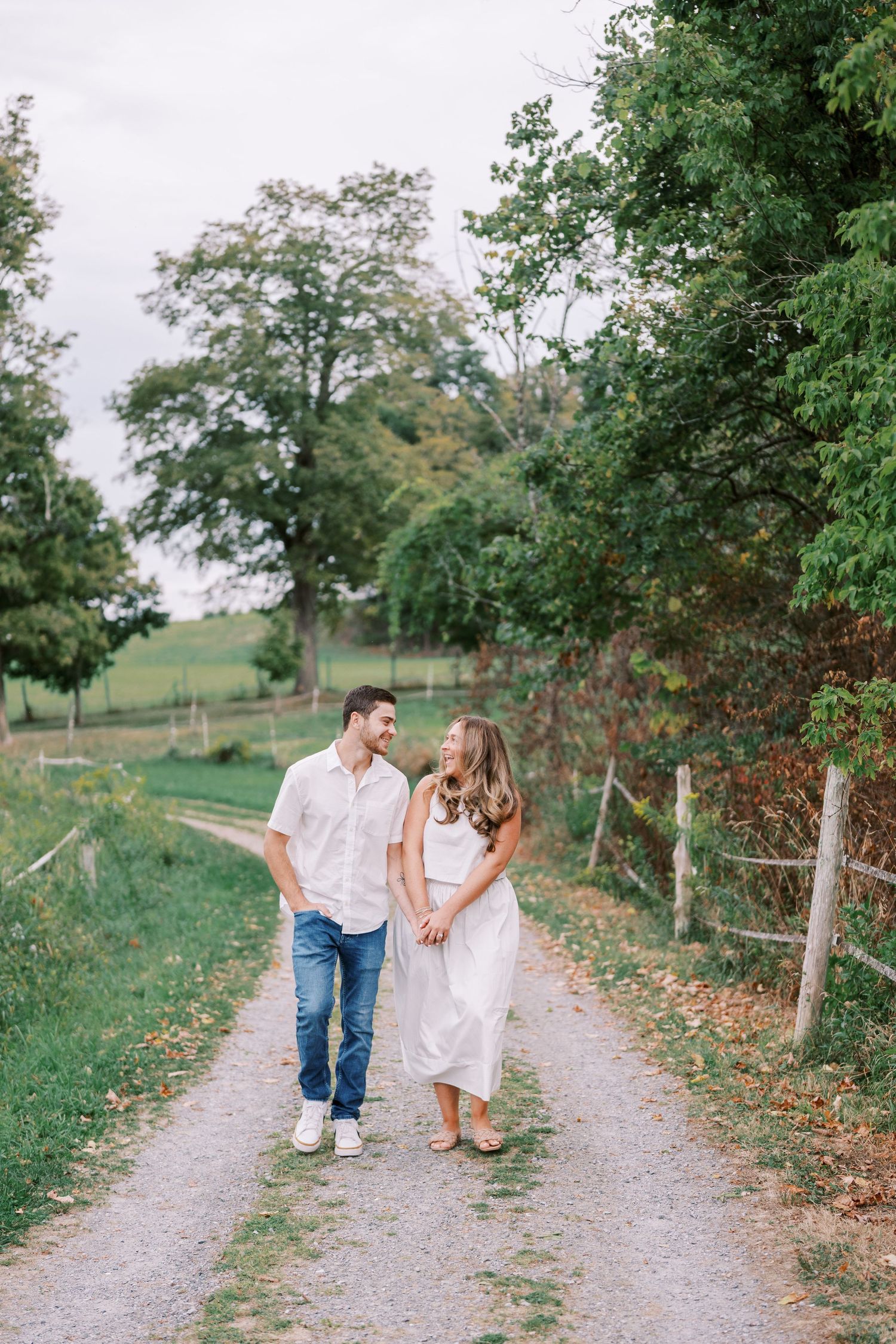 A romantic couple walking hand in hand down a tree-lined gravel path on a summer day.
