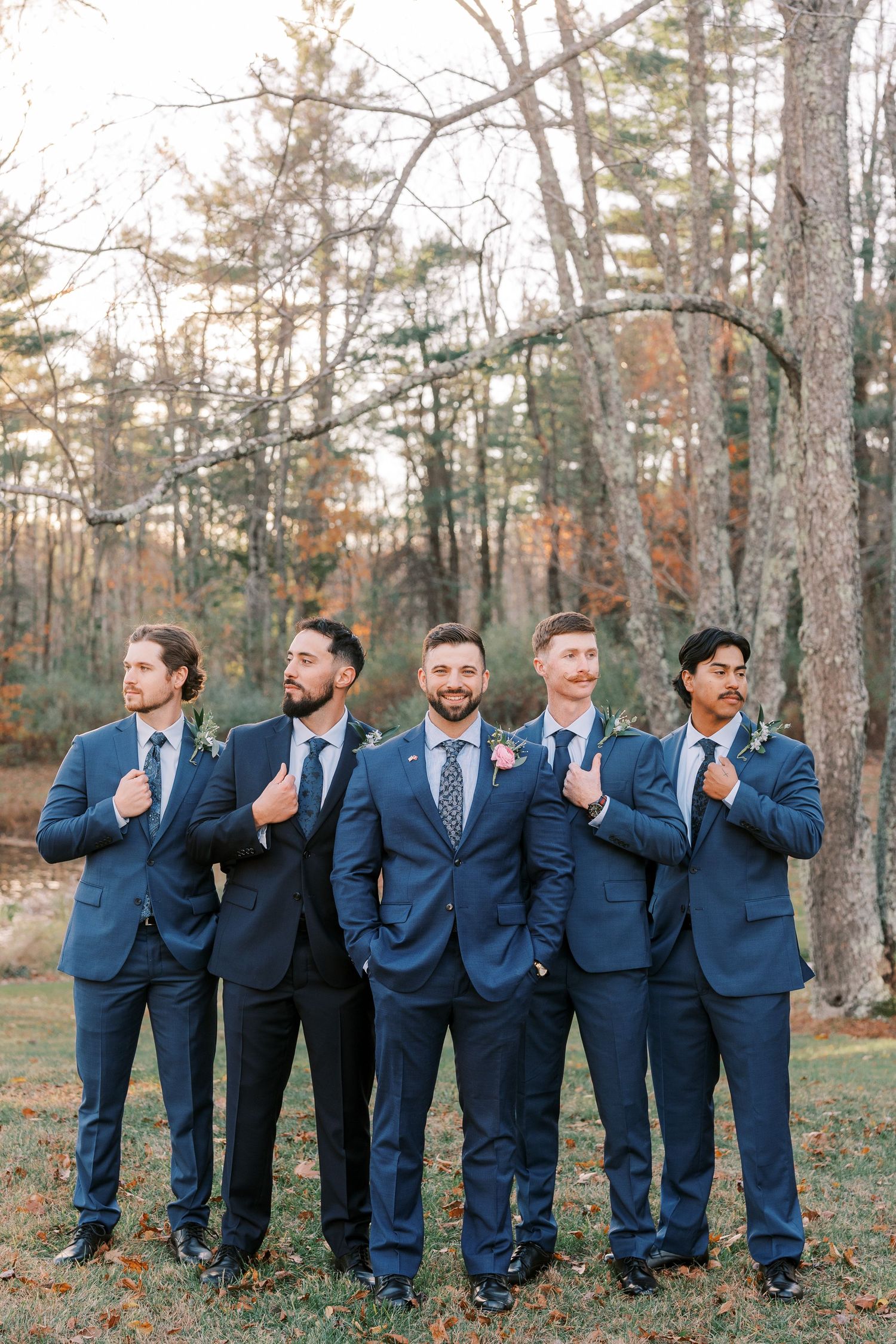 Groomsmen in matching navy blue suits pose together outdoors in a wooded setting during autumn.