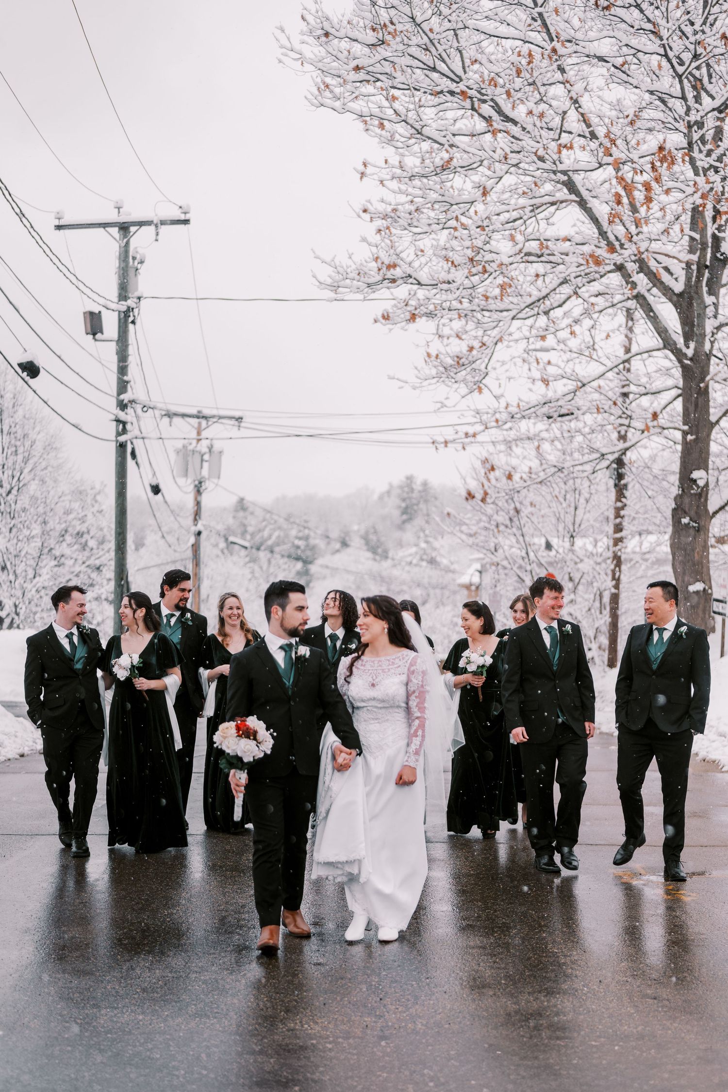 Wedding party walking together on snowy street with bare trees and power lines in background.