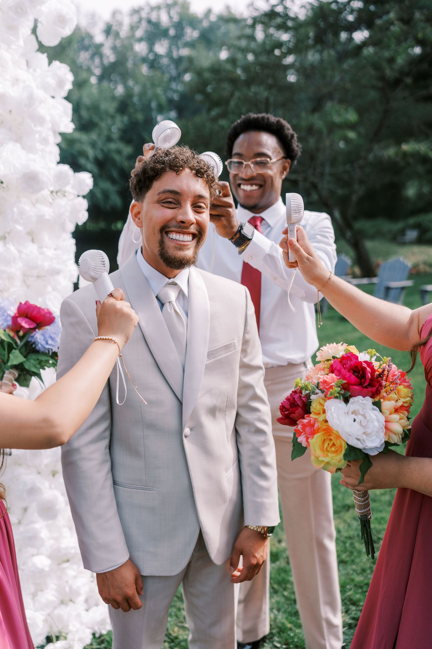 Wedding celebration under a white floral arch with joyful guests and vibrant bouquets of pink and orange flowers.
