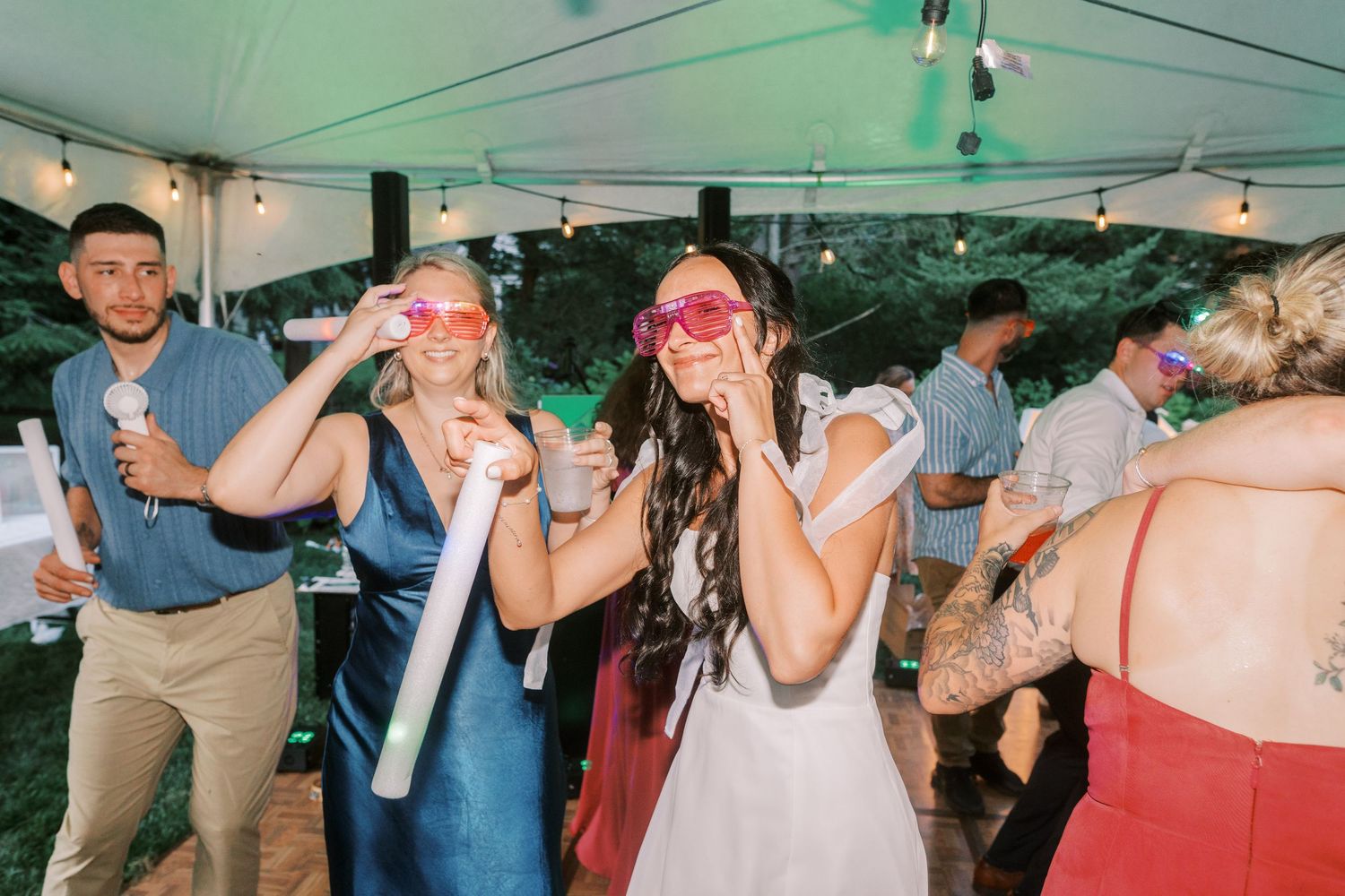 Wedding guests having fun with silly props under a white tent during outdoor reception celebration.