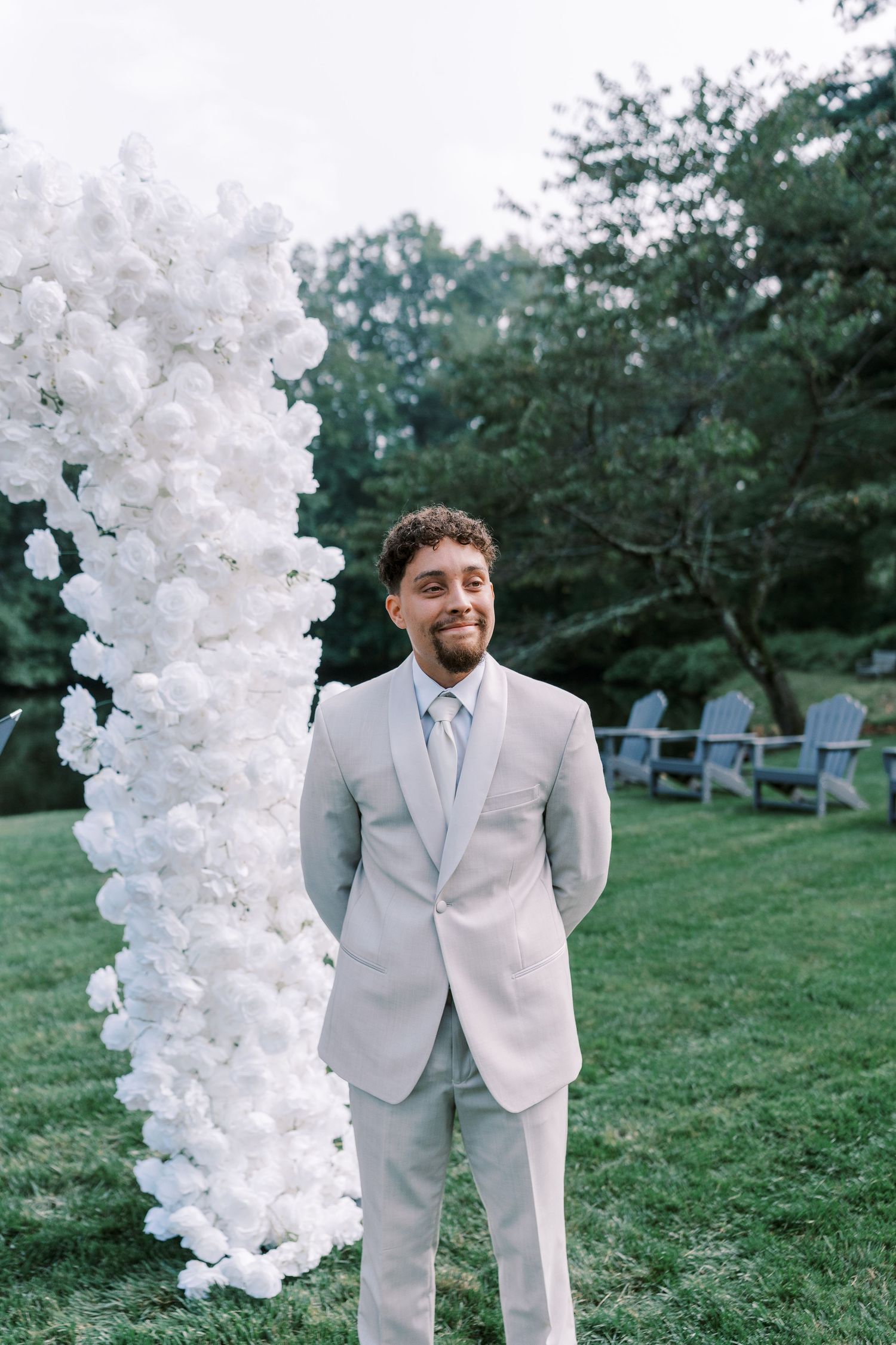 Groom stands next to white floral arch installation in outdoor garden setting.