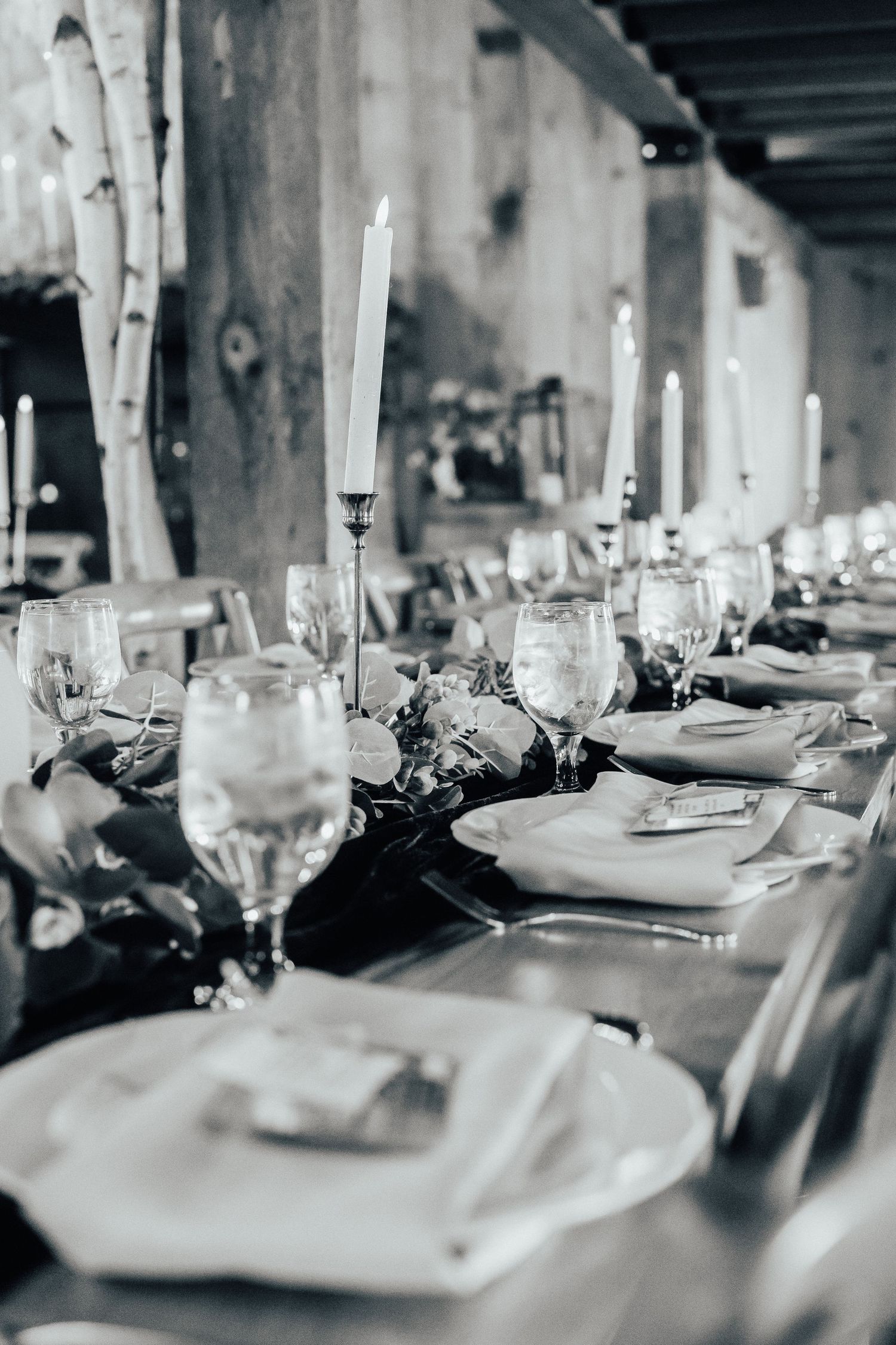Elegant table setting with tall white candles, crystal glasses and formal place settings in black and white tones.