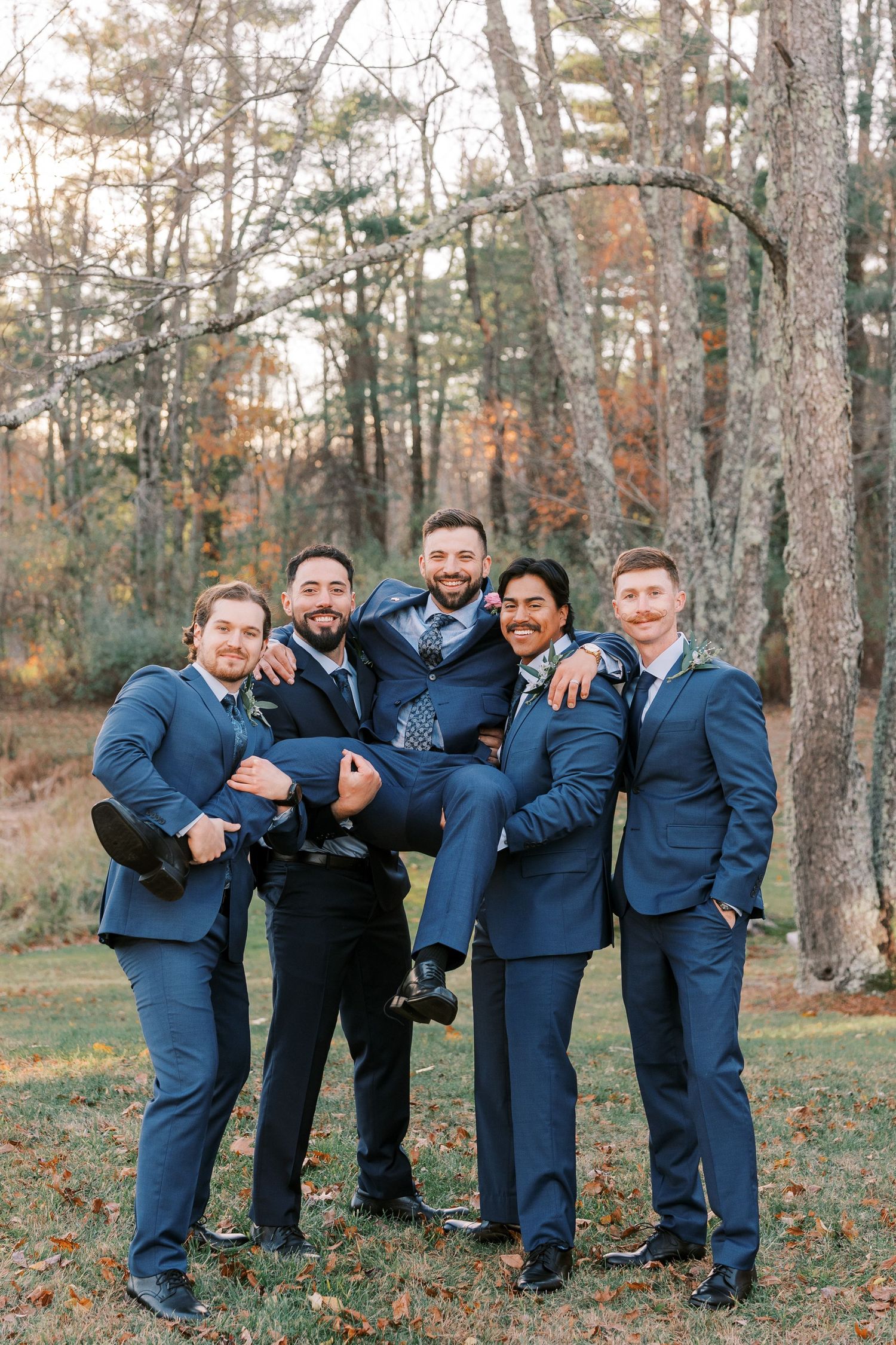A group of groomsmen in navy blue suits playfully pose together in an autumn forest setting.