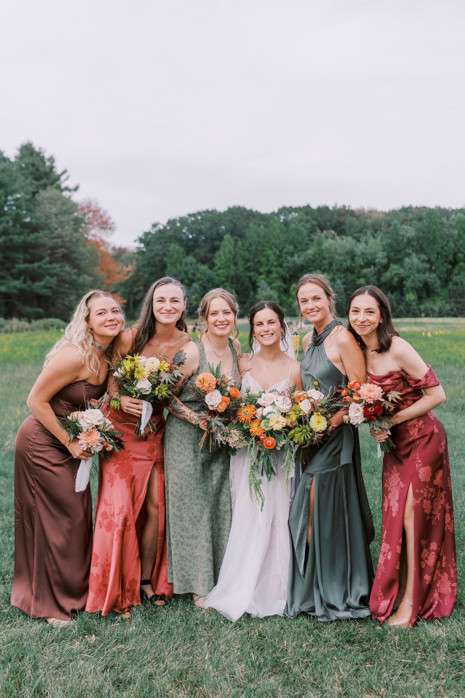 A wedding party in autumn-toned dresses poses together in a grassy field with colorful bouquets.