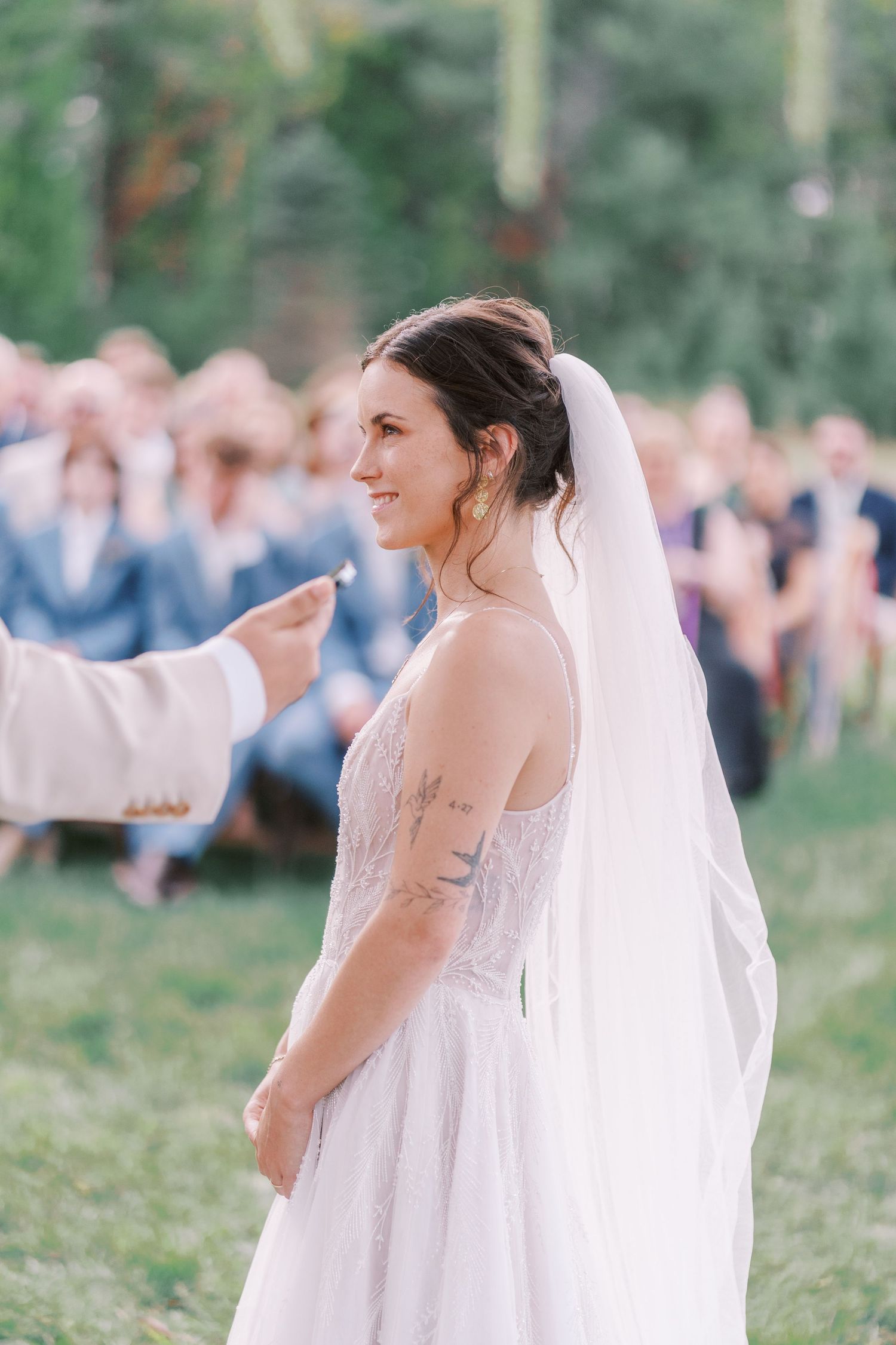 Bride in lace gown with veil poses against natural outdoor backdrop.