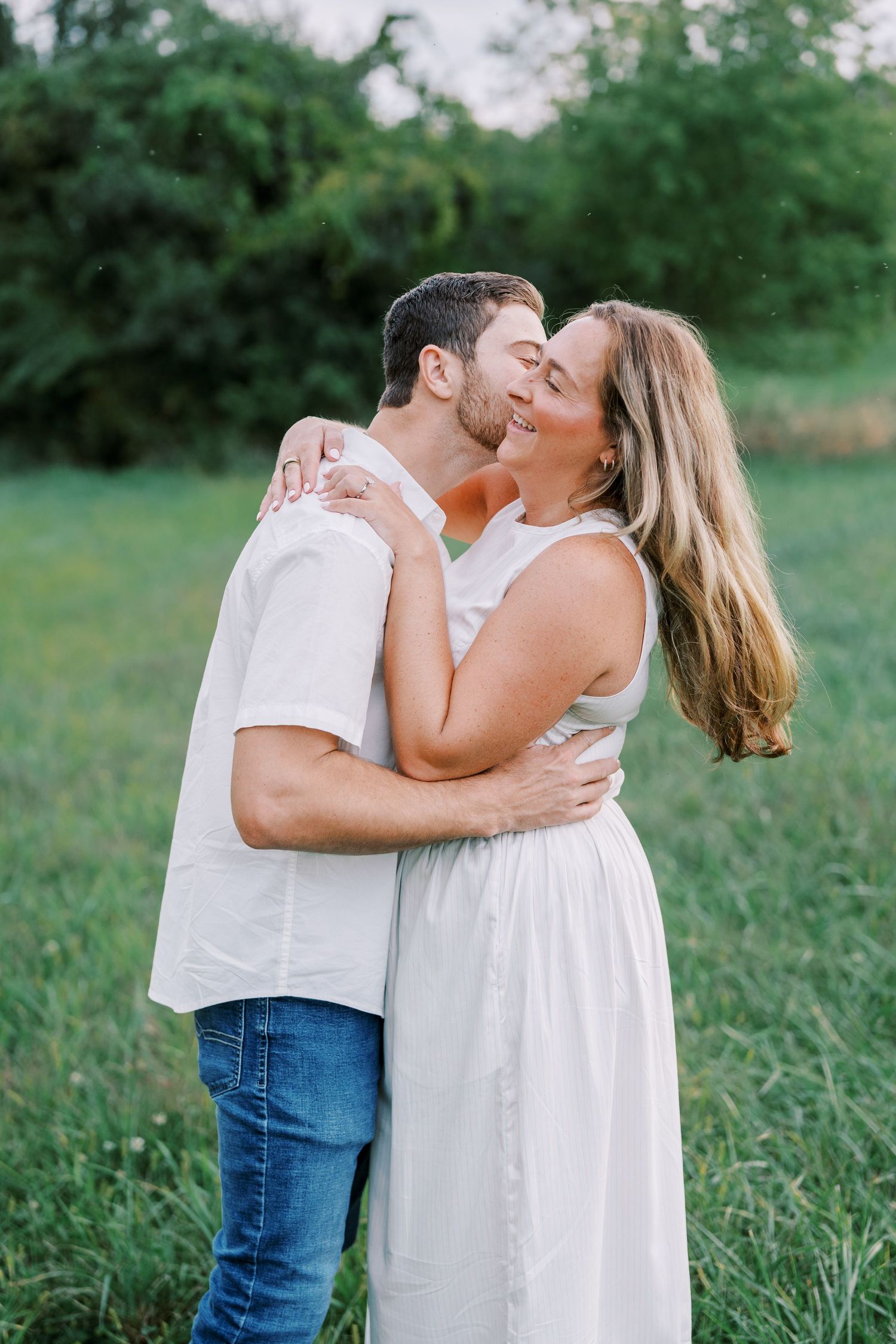 A romantic couple in white outfits sharing a tender kiss while embracing in a lush green outdoor setting.