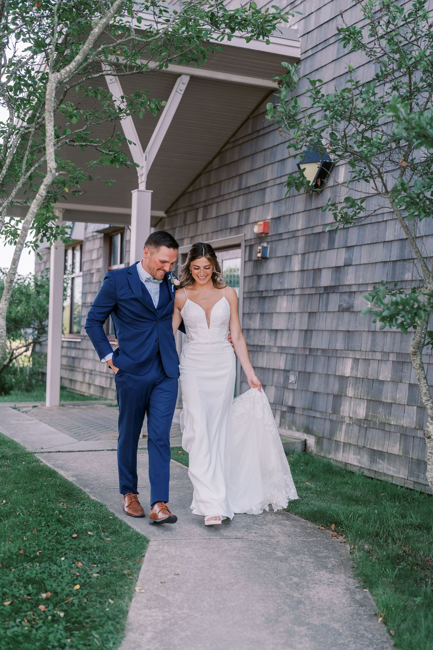 A couple in wedding attire playfully walking and interacting along a rustic building's exterior wall.