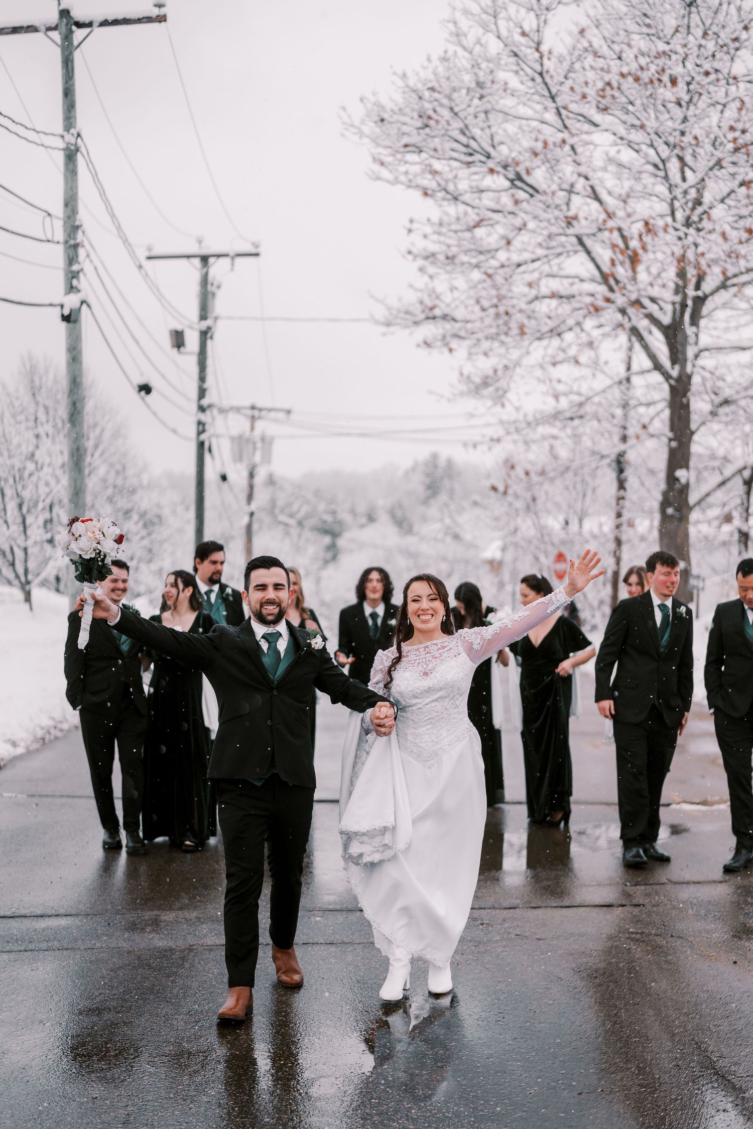 Wedding party in black suits and white dresses poses together on a snowy winter day with bare trees in the background.