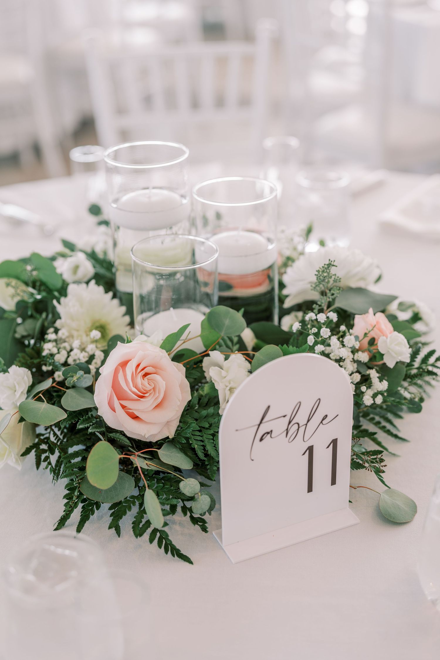 Elegant wedding table centerpiece with blush pink roses, white flowers, greenery and a table number card on white linens.