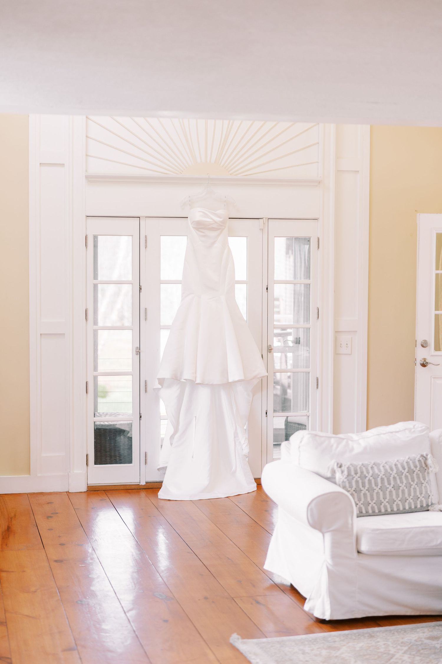 A white wedding dress hangs elegantly in front of French doors in a bright room with hardwood floors and a white couch.