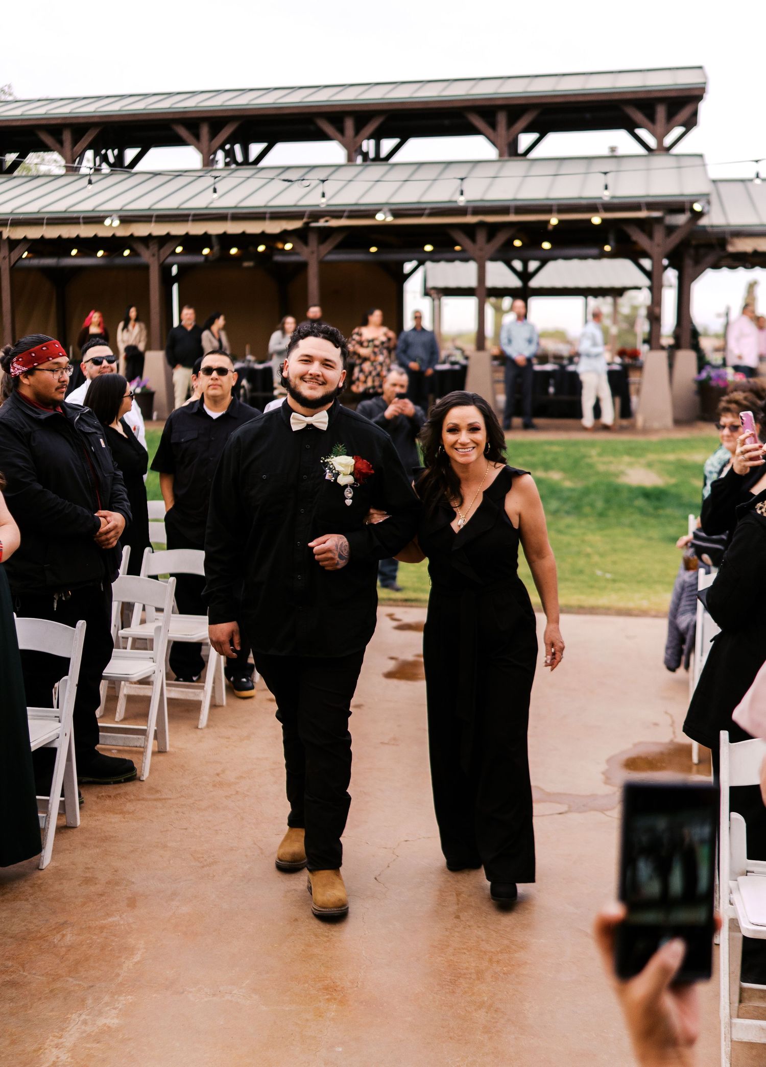 Wedding party members walk down an aisle during an outdoor ceremony with decorative pavilion and string lights.