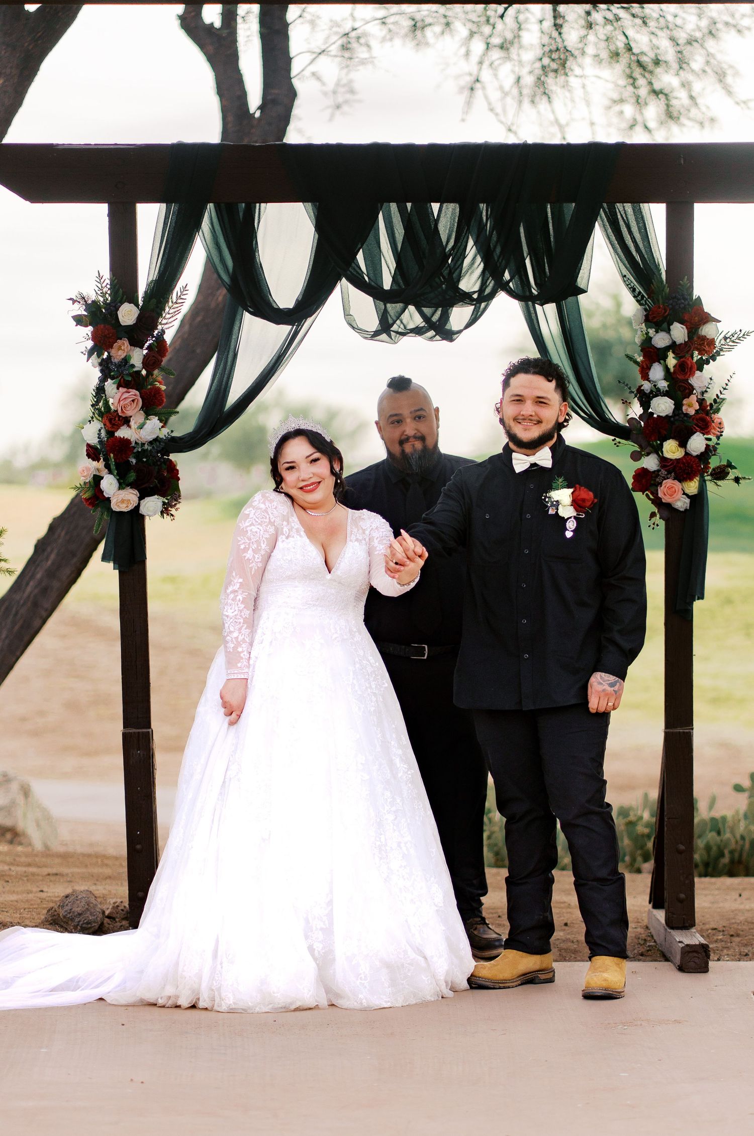 Outdoor wedding ceremony under draped green fabric arch decorated with pink and white flowers at corners.