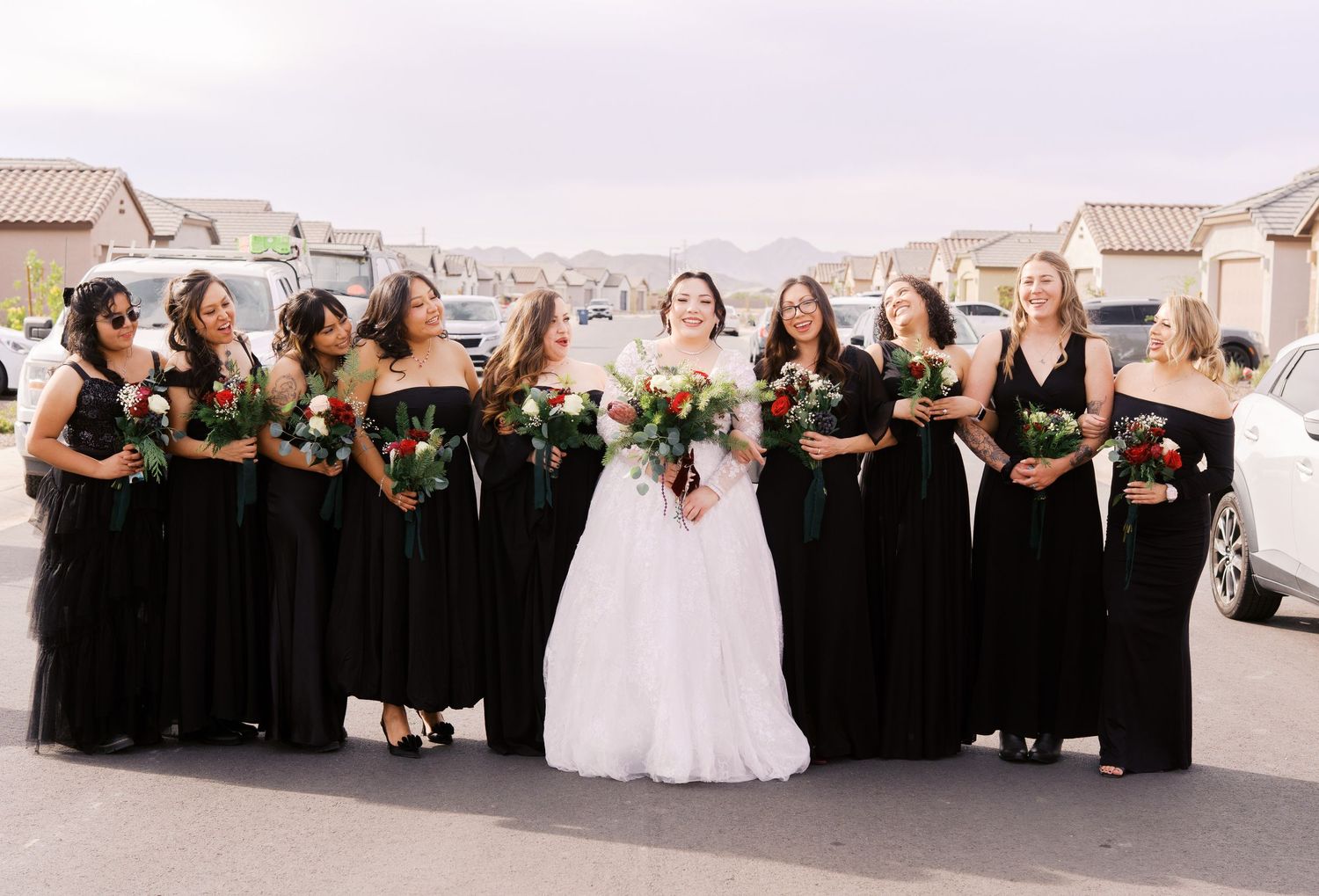 A wedding party poses outdoors wearing black bridesmaid dresses and holding white floral bouquets with the bride in white.