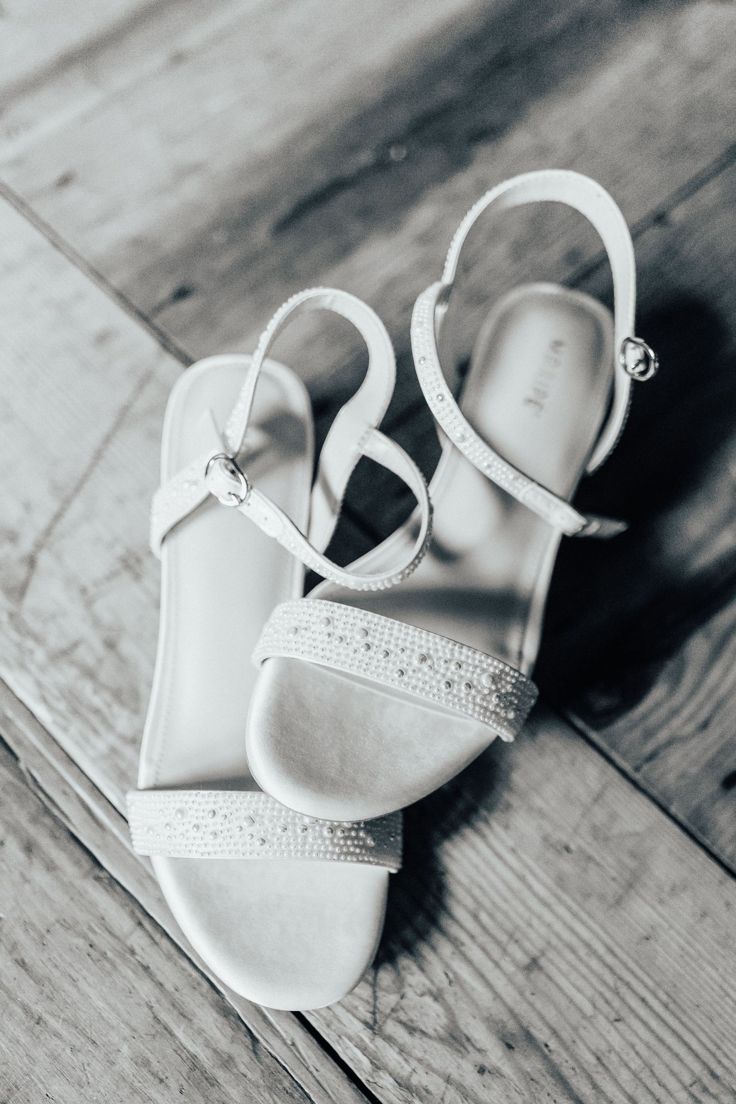 Elegant white bridal sandals with rhinestone straps and ankle buckle closure displayed on wooden floor.