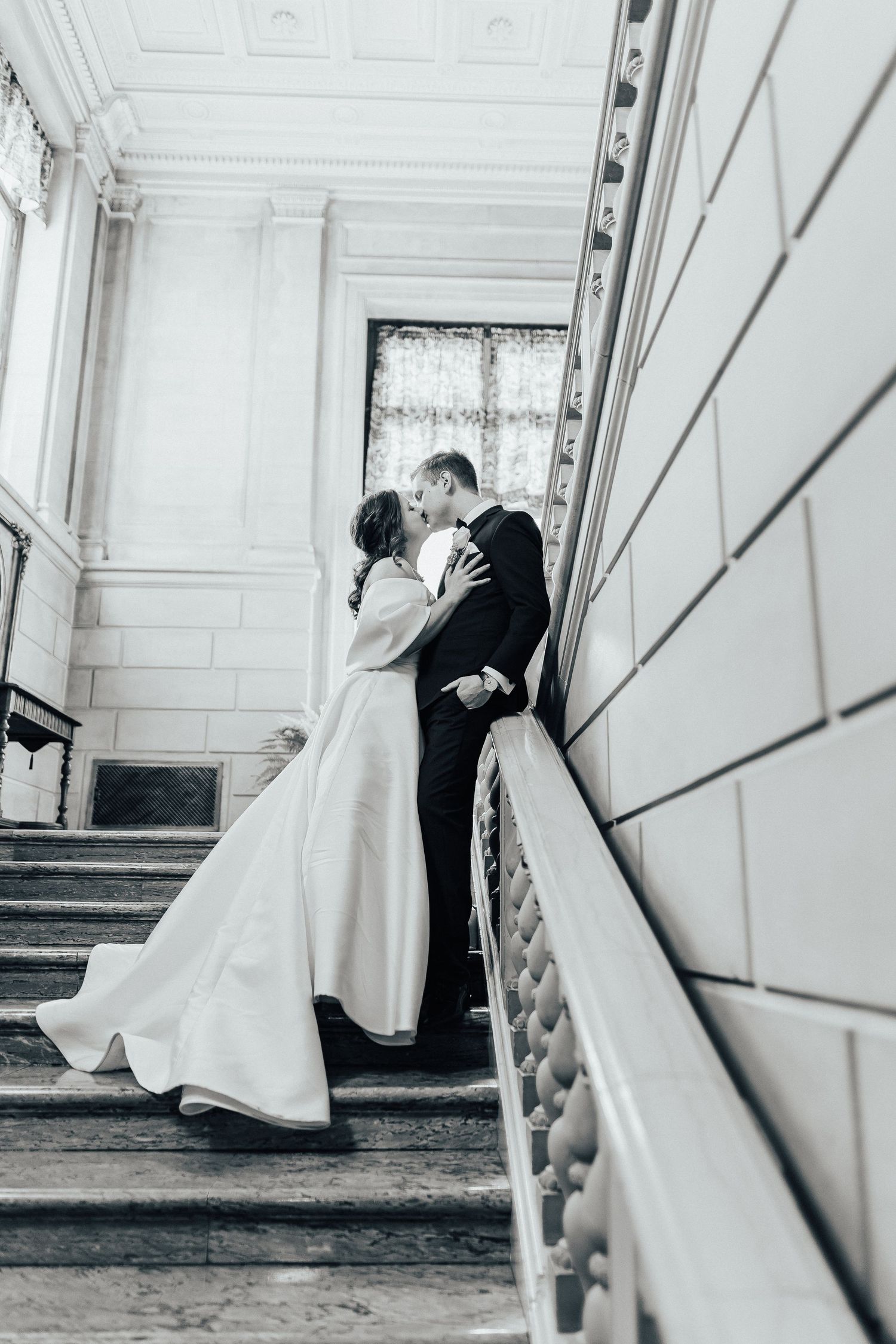 Black and white romantic wedding photo of newlyweds sharing a kiss on elegant mansion staircase.