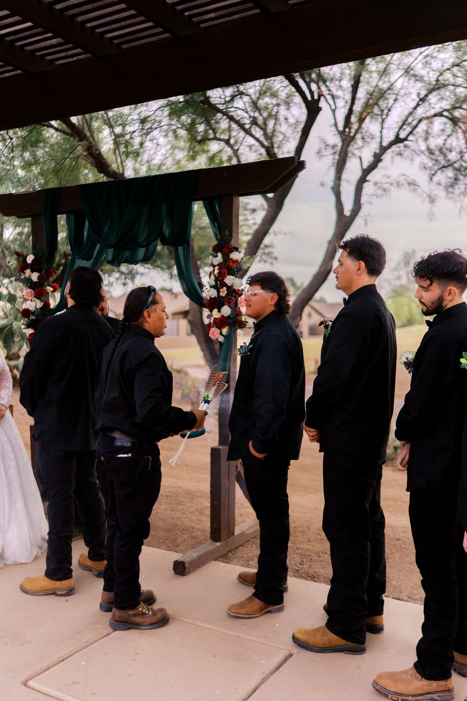 A formal wedding party in matching black outfits stand together under a covered outdoor ceremony venue.