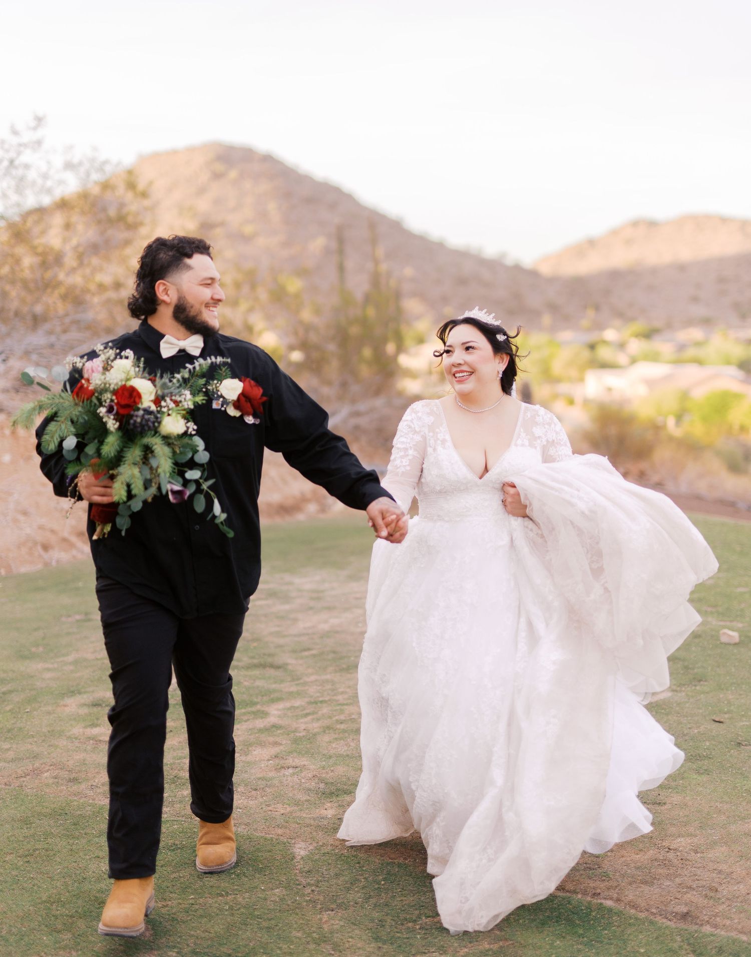 A couple in wedding attire holding hands and laughing while walking on grass with desert mountains in background.