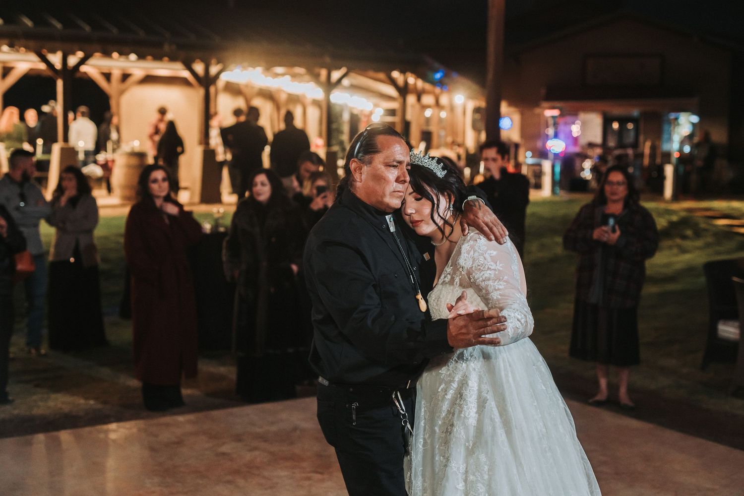 A couple shares their first dance at an outdoor evening wedding reception under a wooden pavilion as guests watch.