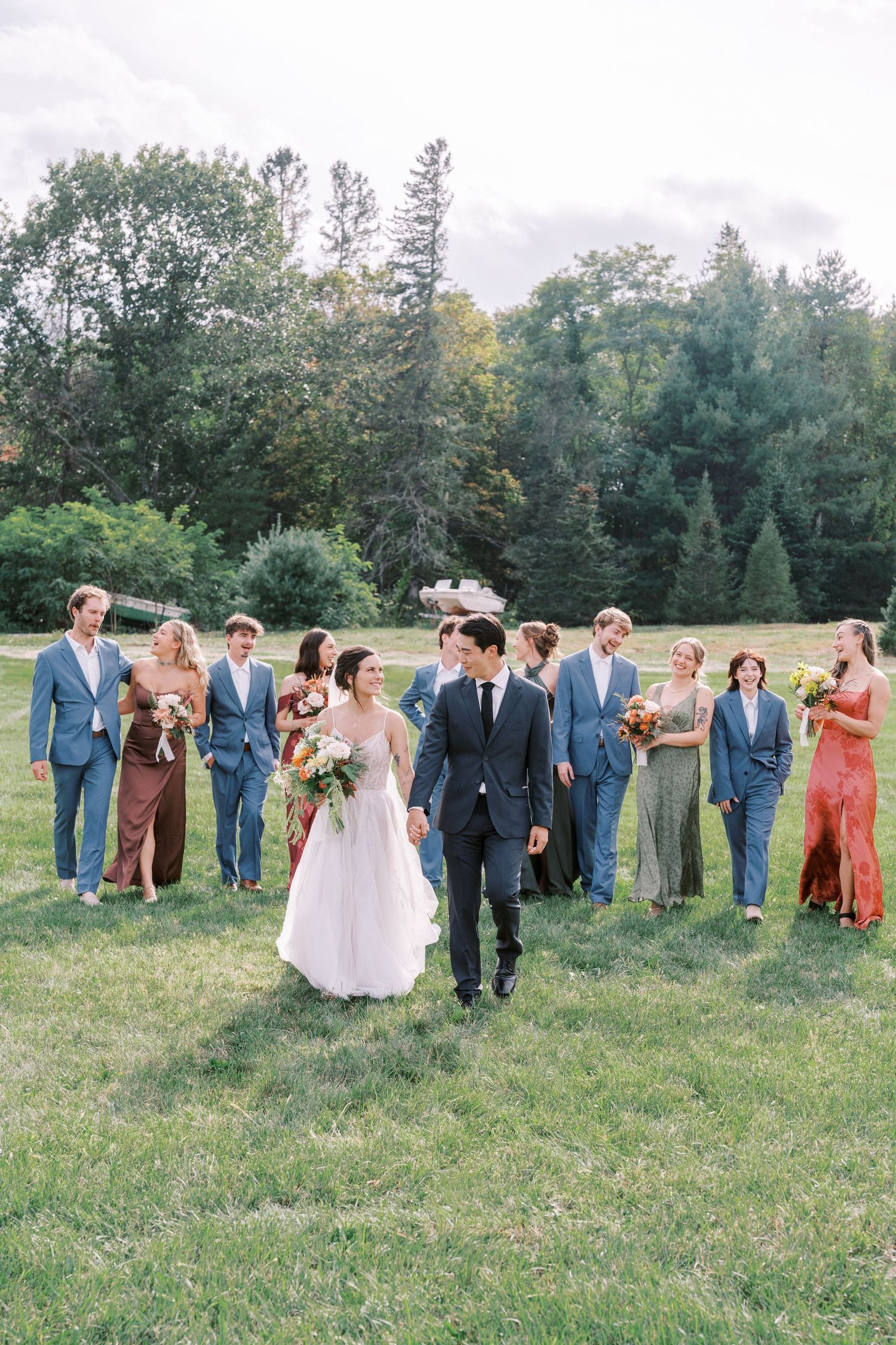 Wedding party in blue suits and coral dresses poses together on grassy field with trees in background.