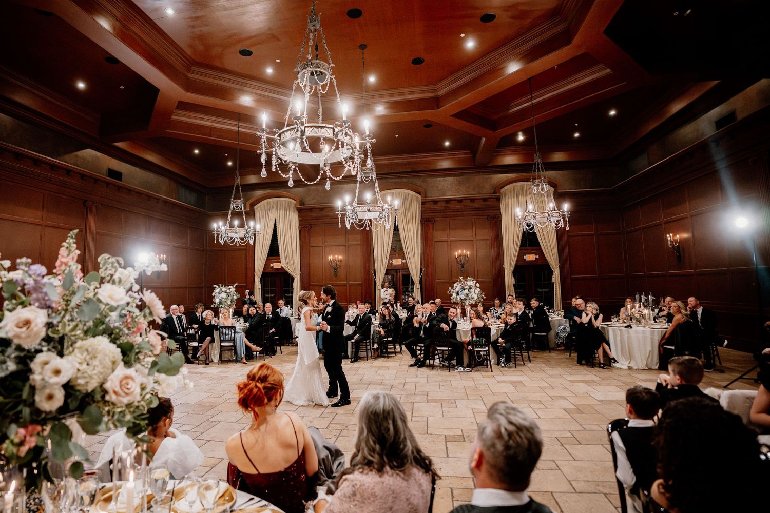 A panoramic view of an elegant ballroom wedding reception with crystal chandeliers and guests dancing on the dance floor.