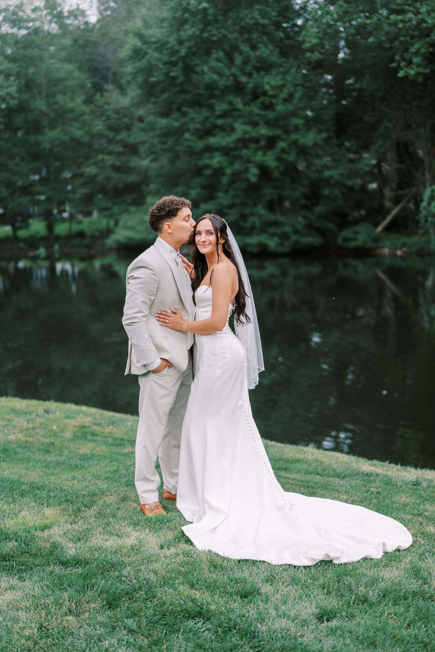 Newlyweds share intimate moment by scenic pond with bride in flowing white gown and groom in light grey suit.