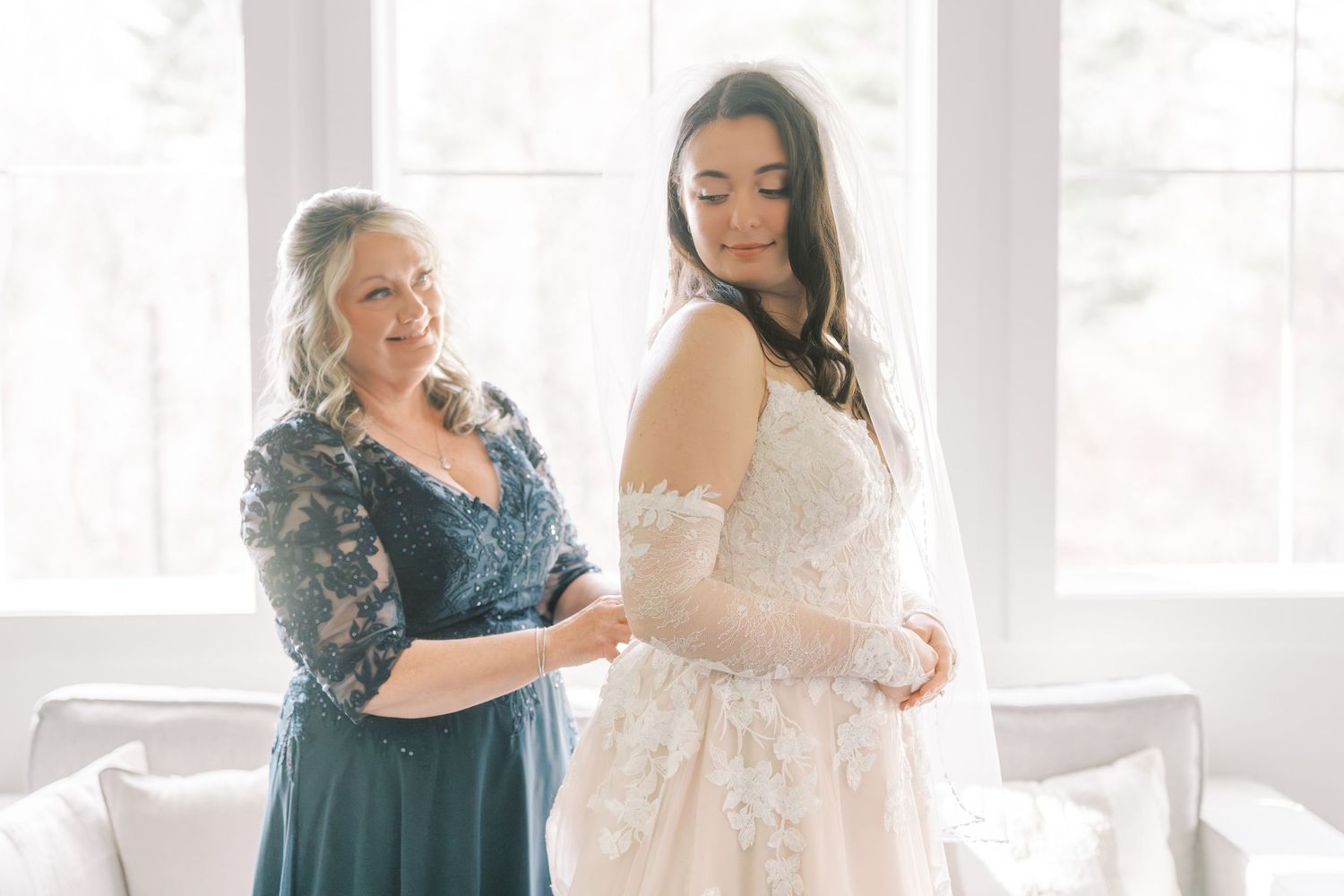 Wedding party members help with final dress preparations in a bright sunlit room.