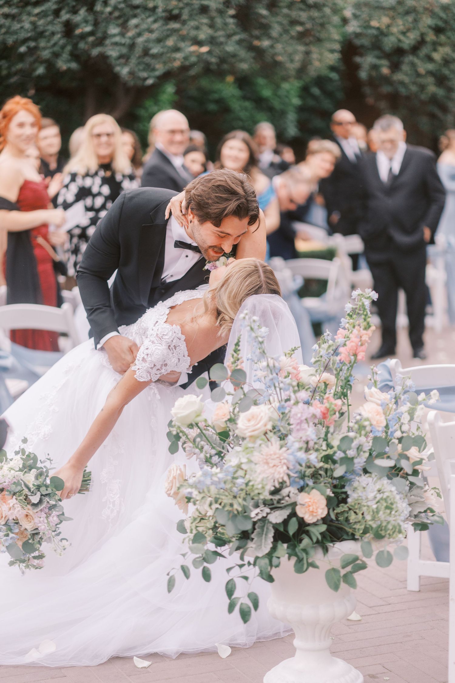 Romantic wedding couple shares intimate dance dips with elegant floral arrangements and guests in outdoor setting.