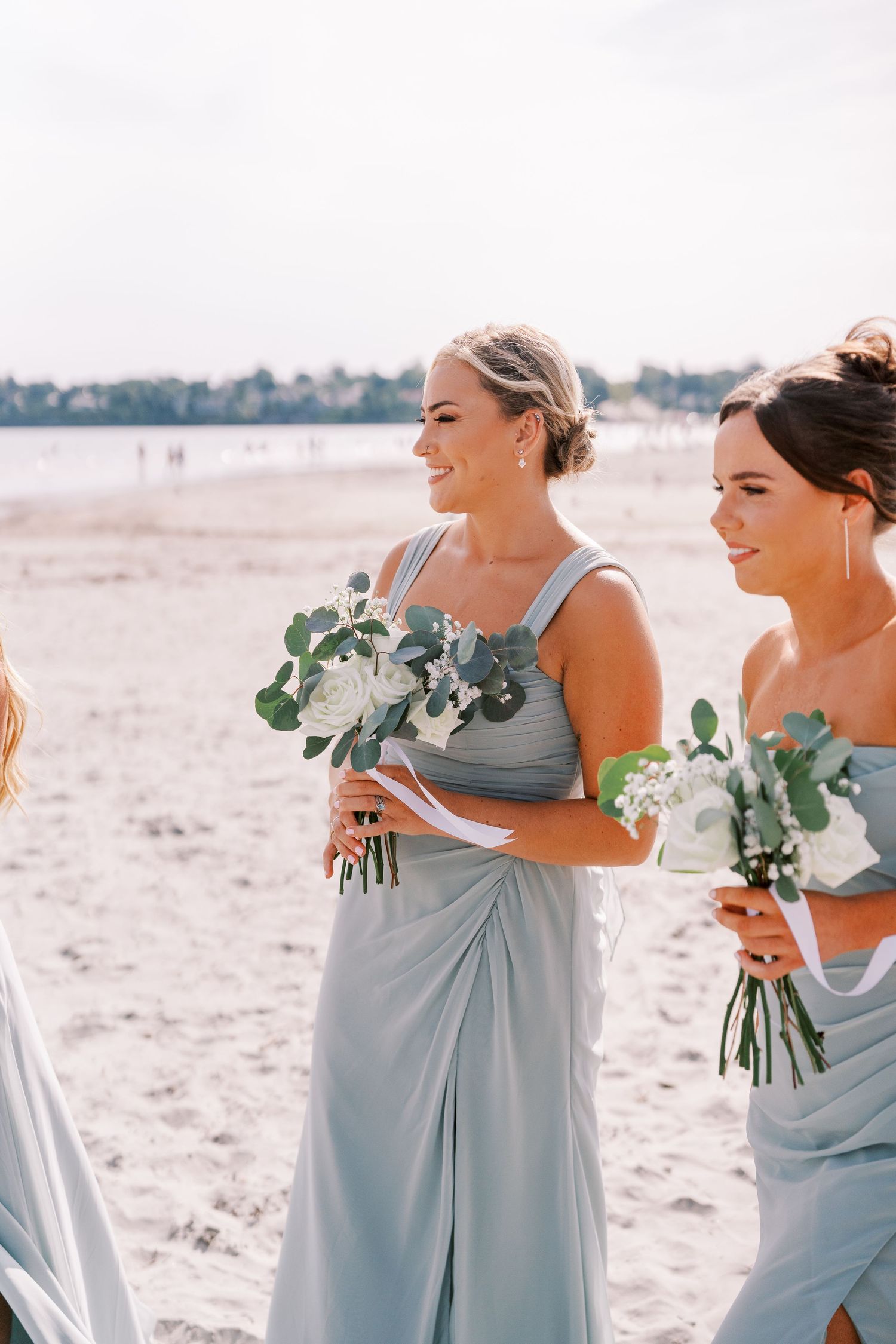 Bridesmaids in pale blue dresses hold eucalyptus bouquets while standing on a sunny beach.