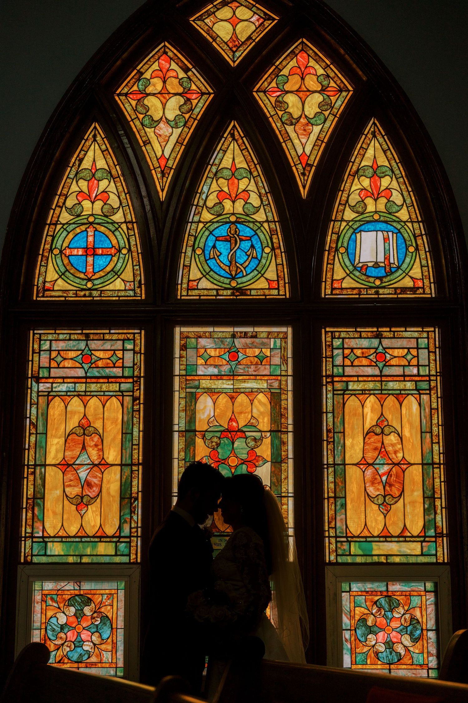 A silhouetted couple stands before a colorful stained glass church window.