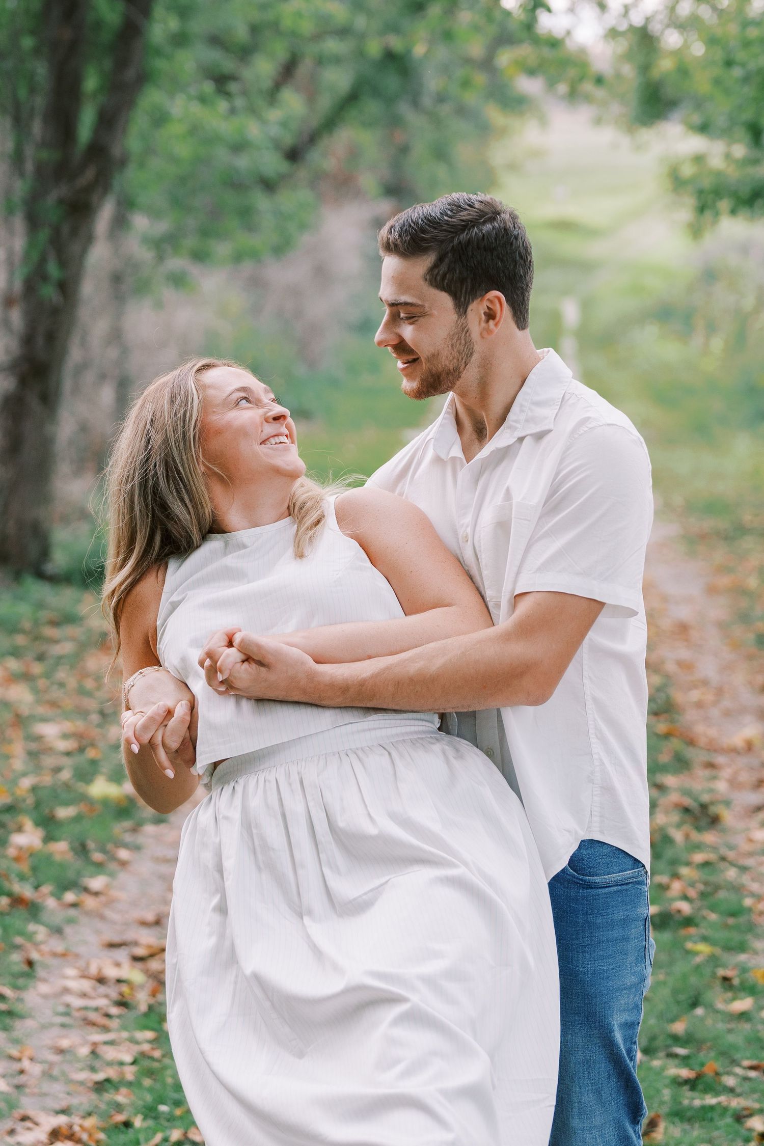 A couple in white clothing dances and embraces joyfully in a natural outdoor setting.