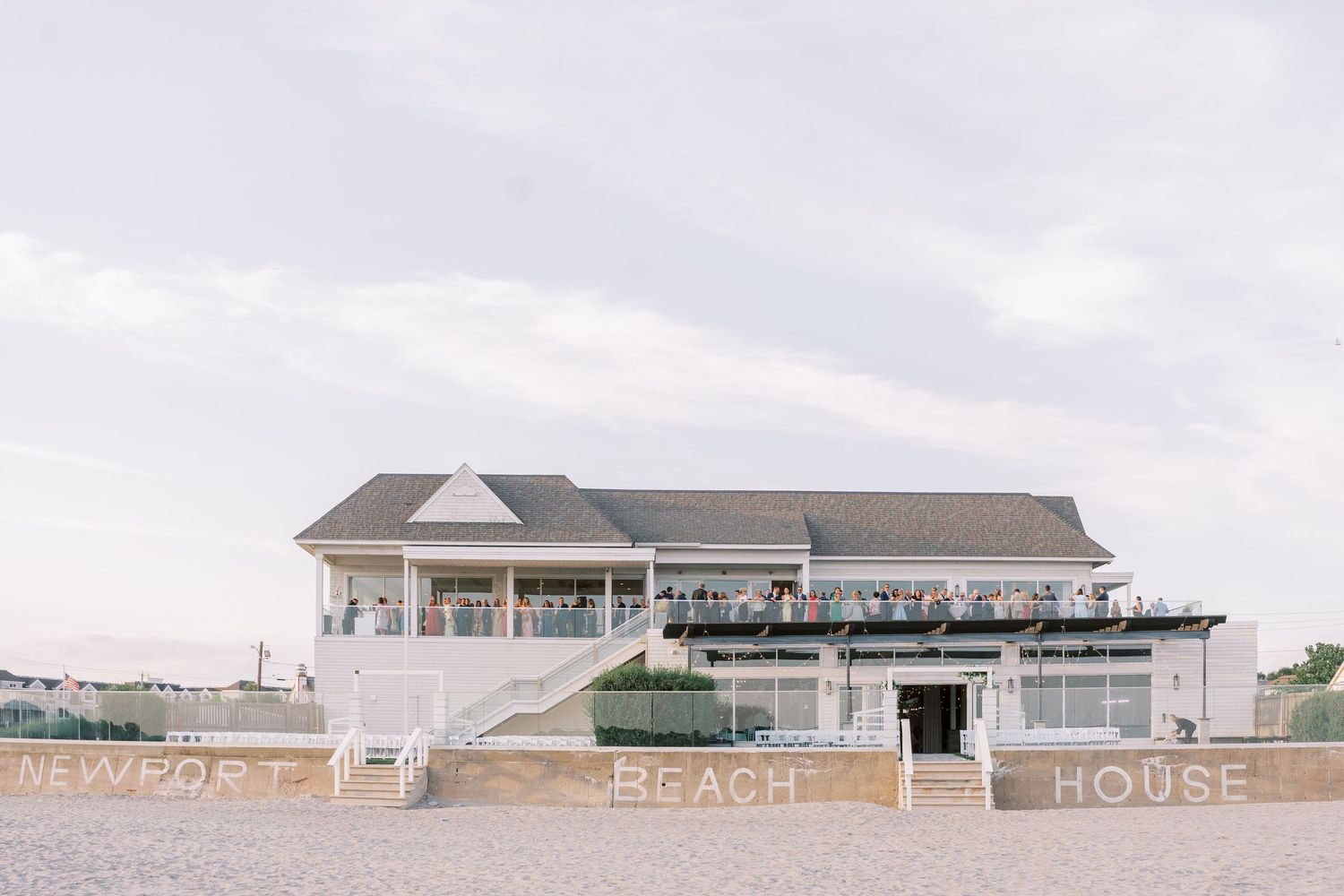 A modern beachfront house with a large deck and 'Newport Beach House' signage on its white exterior.