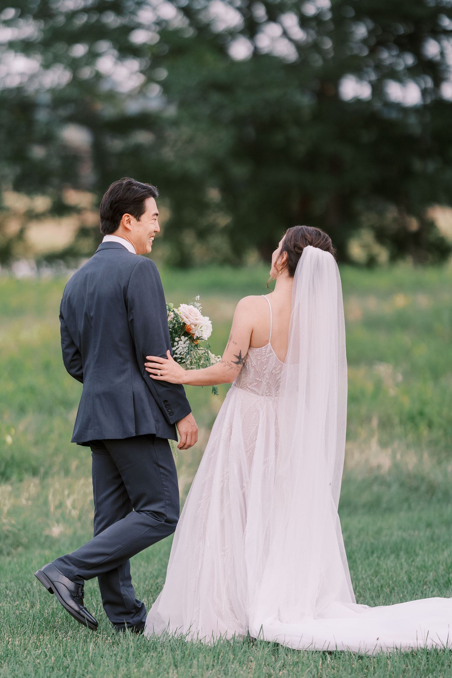Couple walking together across grassy field with flowing white wedding veil.