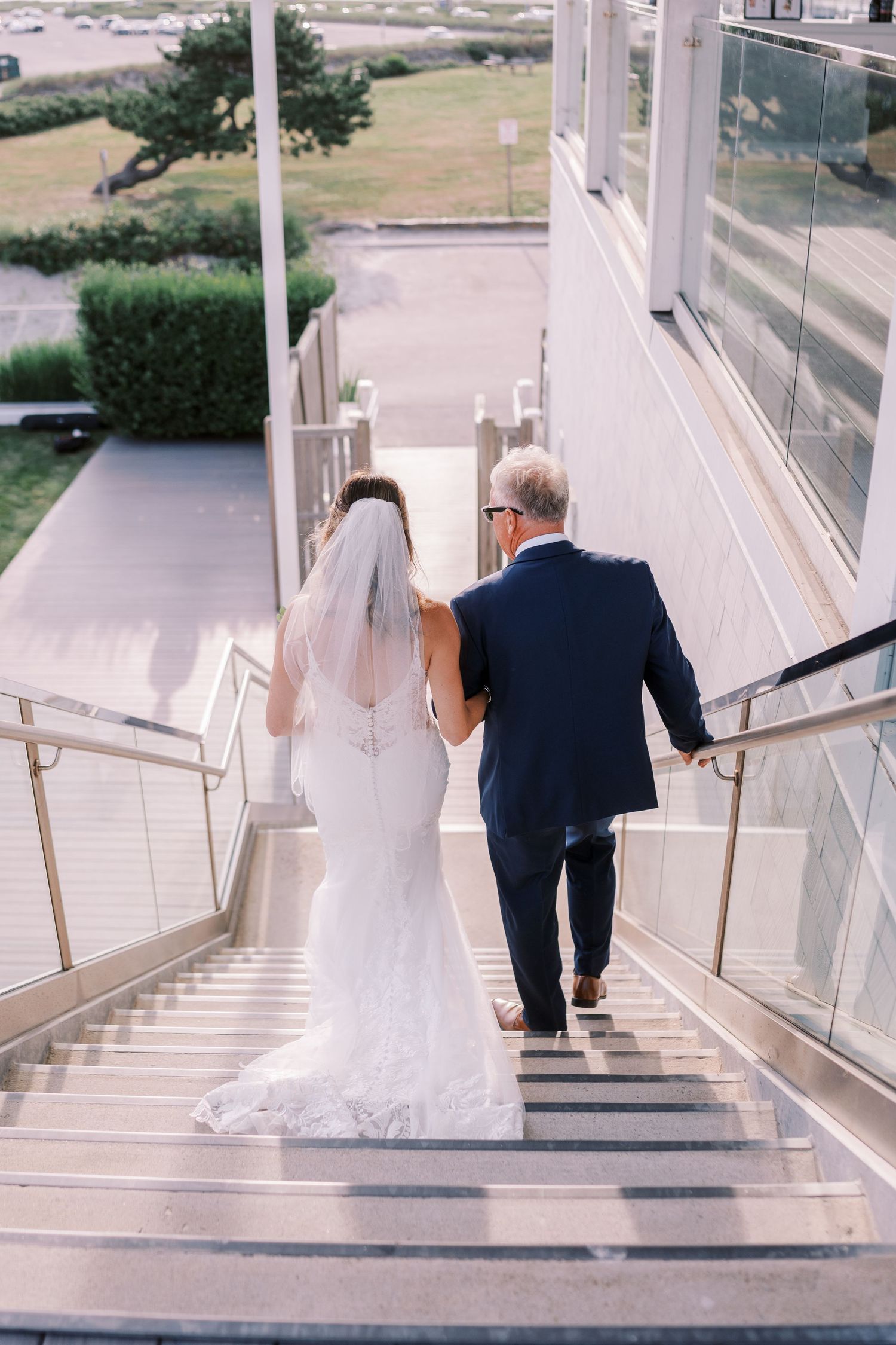 A couple in wedding attire descend modern glass-lined stairs together, with the bride's white veil and gown trailing behind.