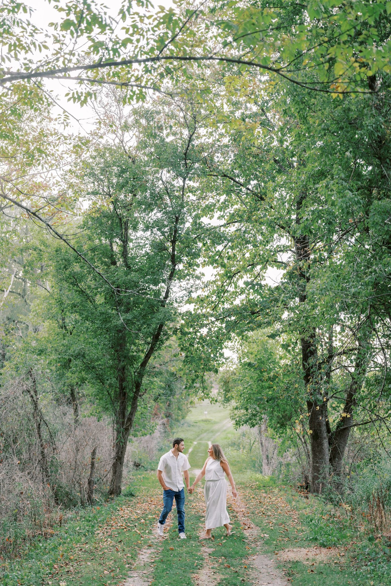 A couple walks hand in hand down a tree-lined path surrounded by lush green foliage.