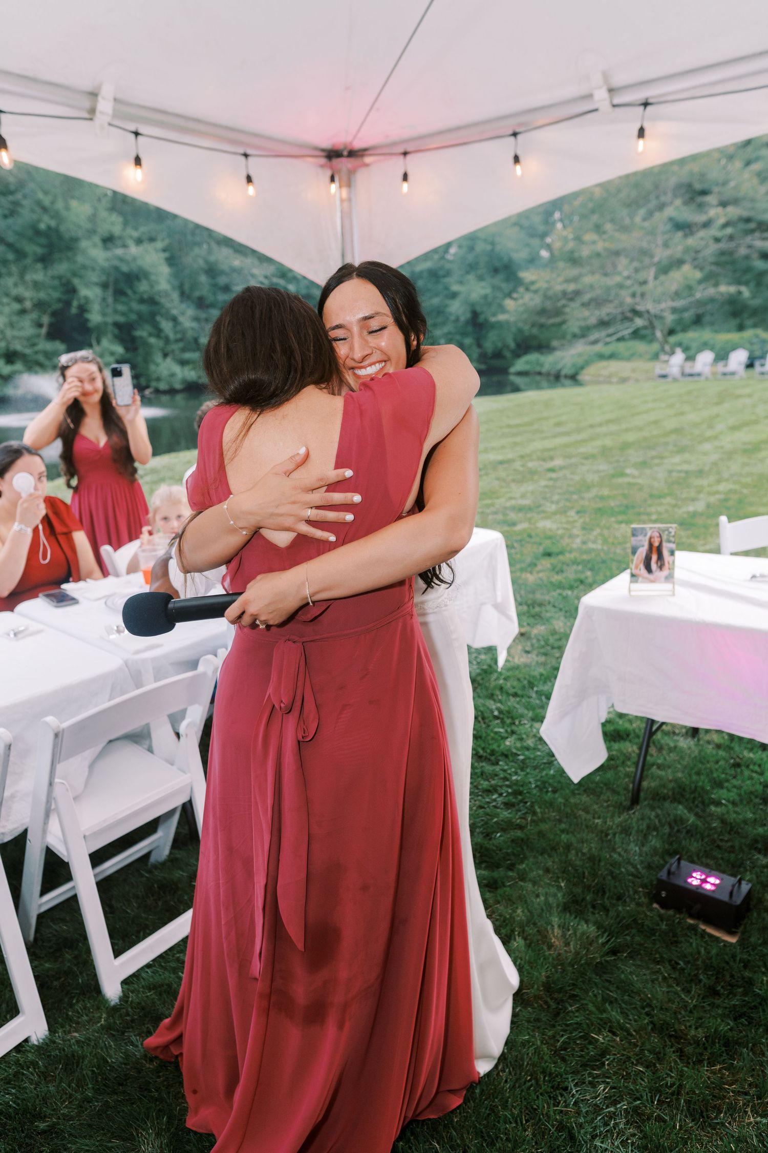 Two people share an emotional embrace at an outdoor wedding reception under a white tent with string lights.