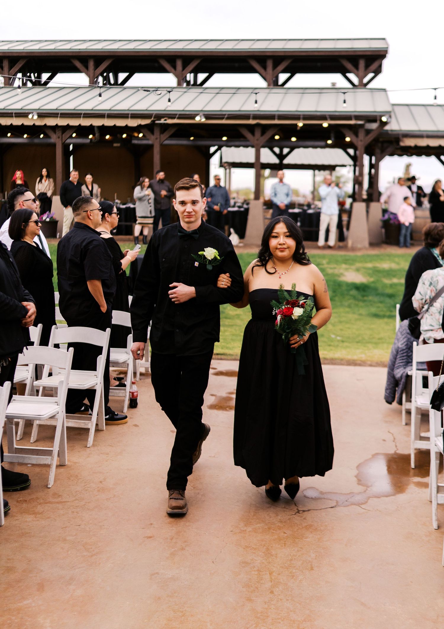 Wedding party members in black formal attire walk down an outdoor aisle during a ceremony at a covered venue.