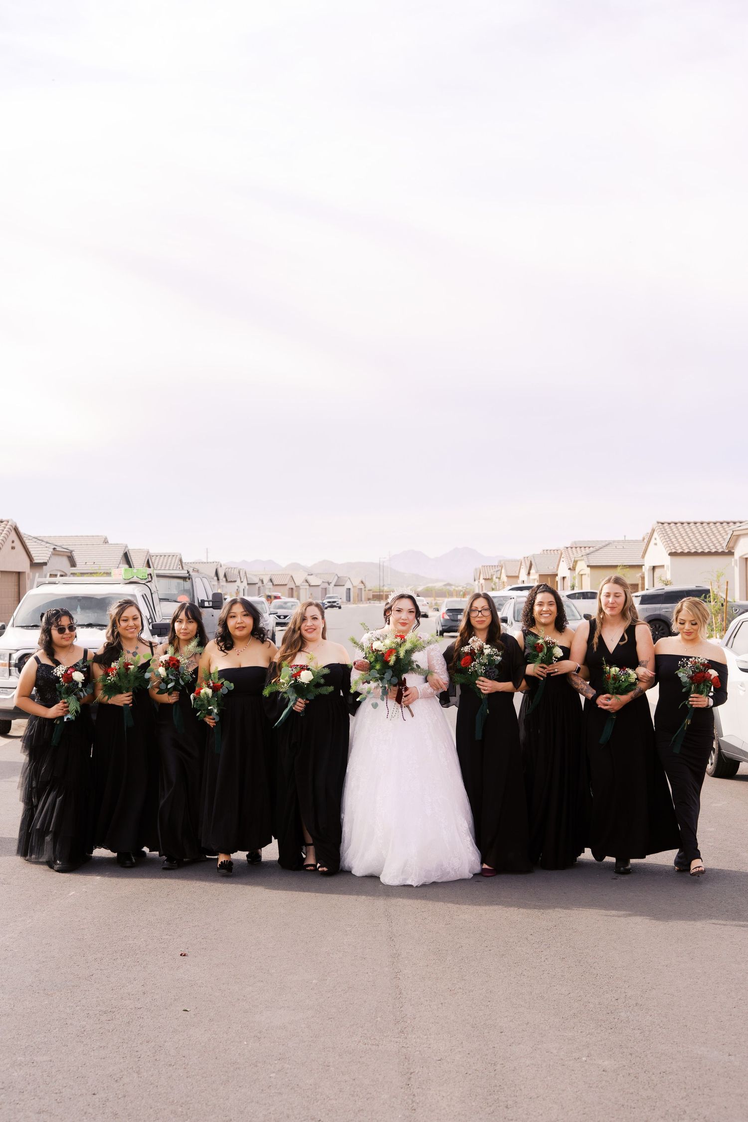A bridal party wearing black bridesmaid dresses and a white wedding gown pose together on a residential street.