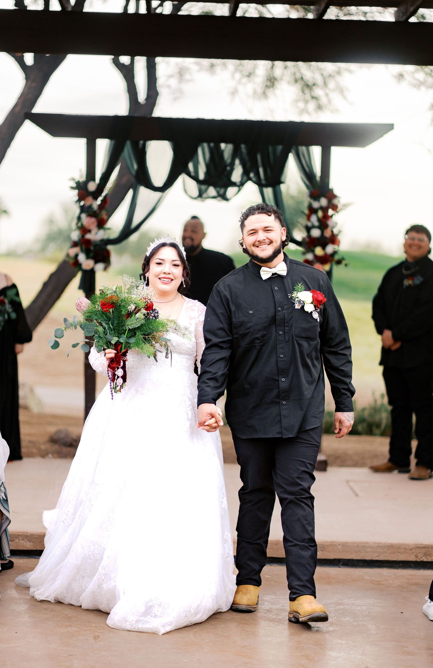 A bride in a white gown and groom in black attire hold hands while walking under a decorated wedding ceremony arch.