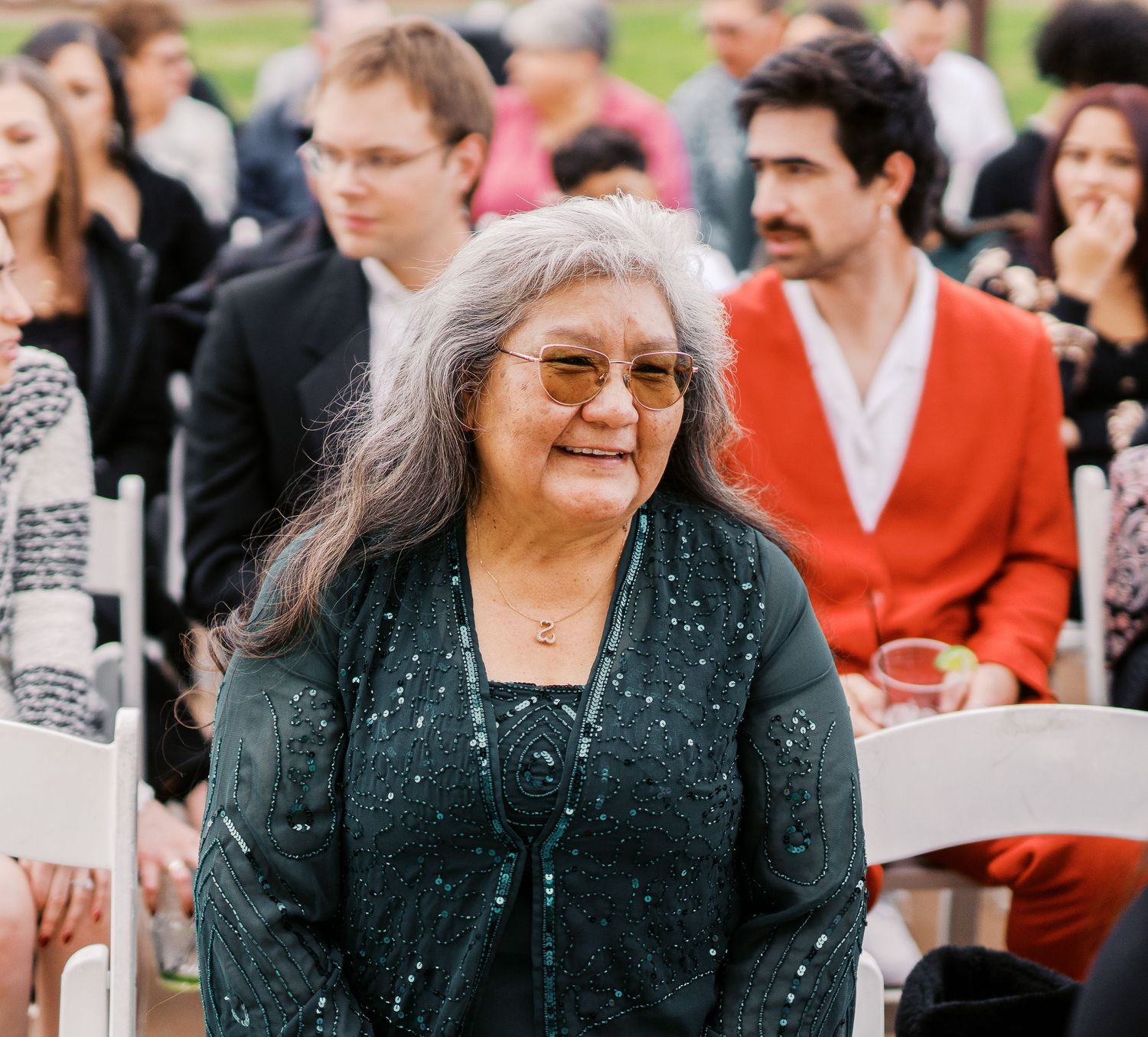 A person with long gray hair and glasses smiles warmly while wearing a dark polka dot top at an outdoor gathering.