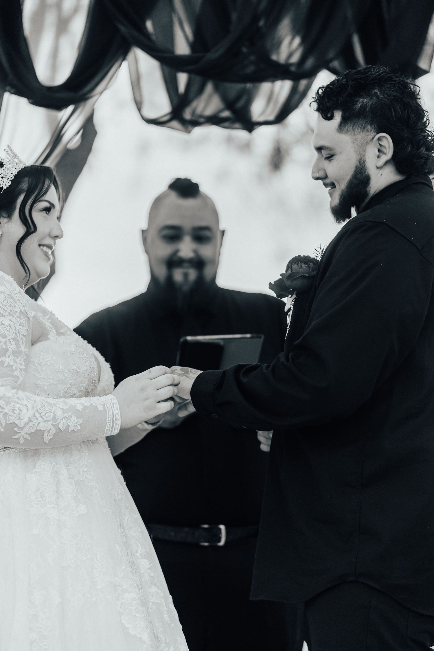 A black and white wedding ceremony photo showing a couple exchanging rings with an officiant standing between them.