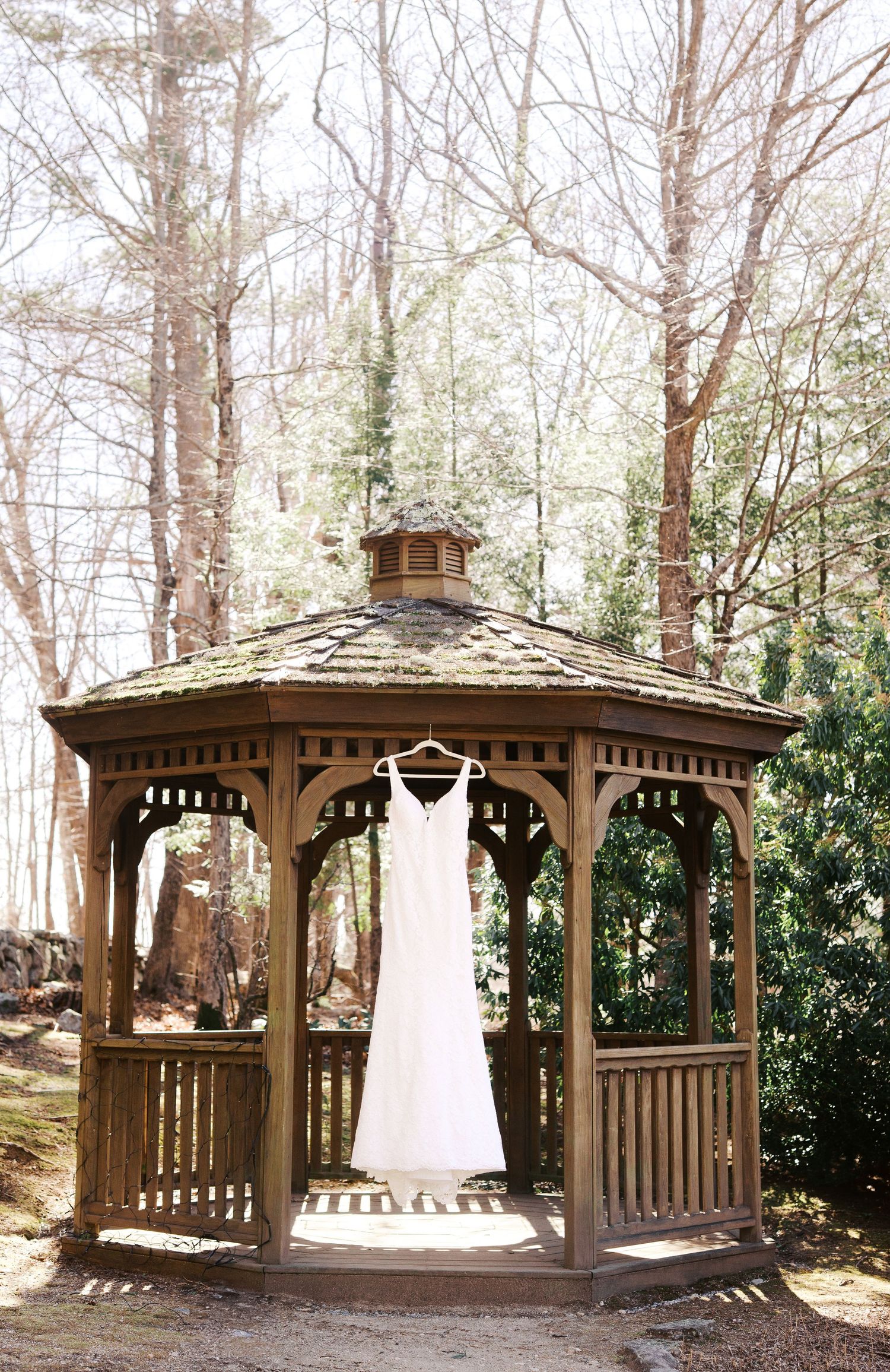 Elegant white wedding dress hanging in a rustic wooden gazebo surrounded by trees in a woodland setting.